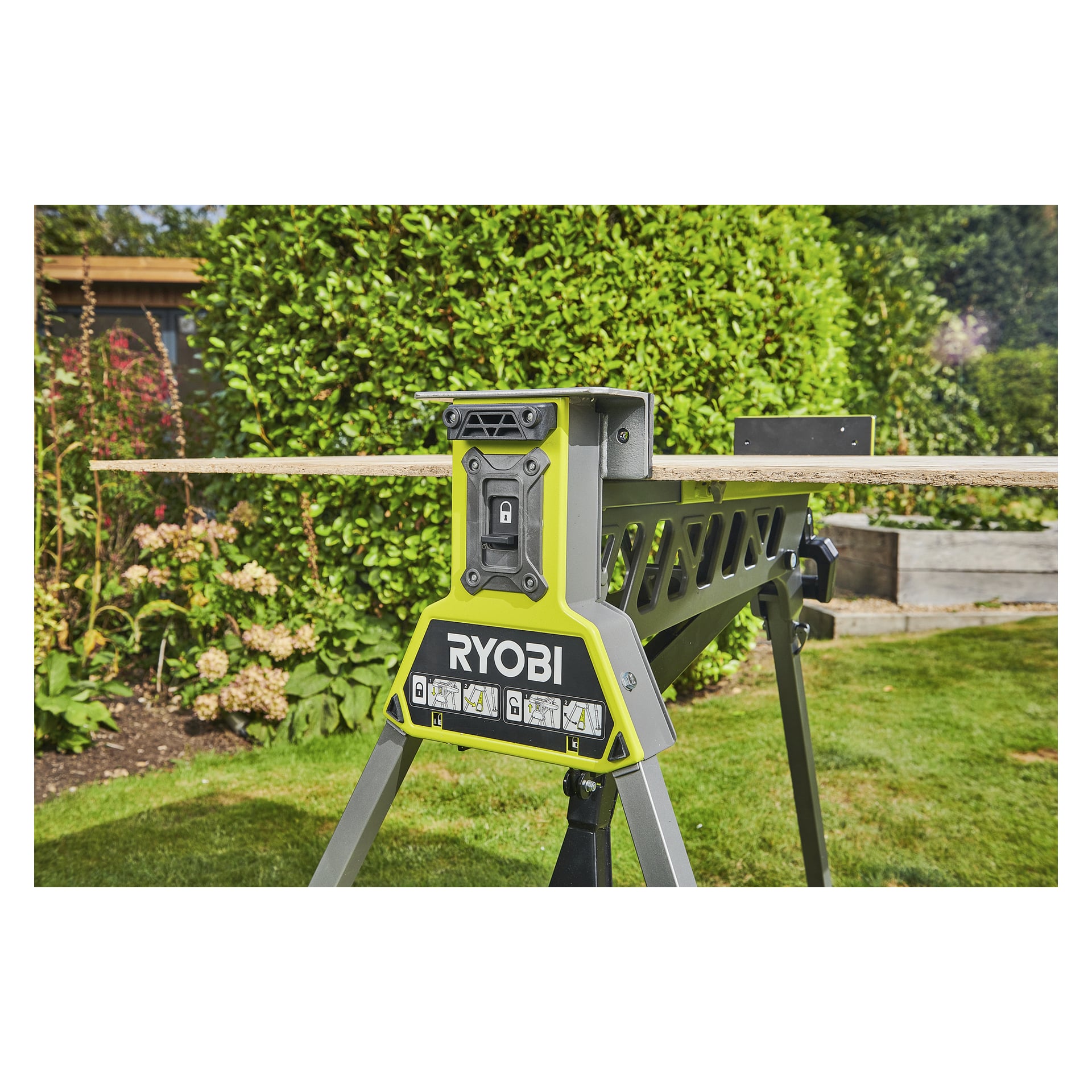 En grön och svart Ryobi One+ fällbar arbetsbänk med klämma håller en träplanka på gräset, med växter och en upphöjd bädd bakom.