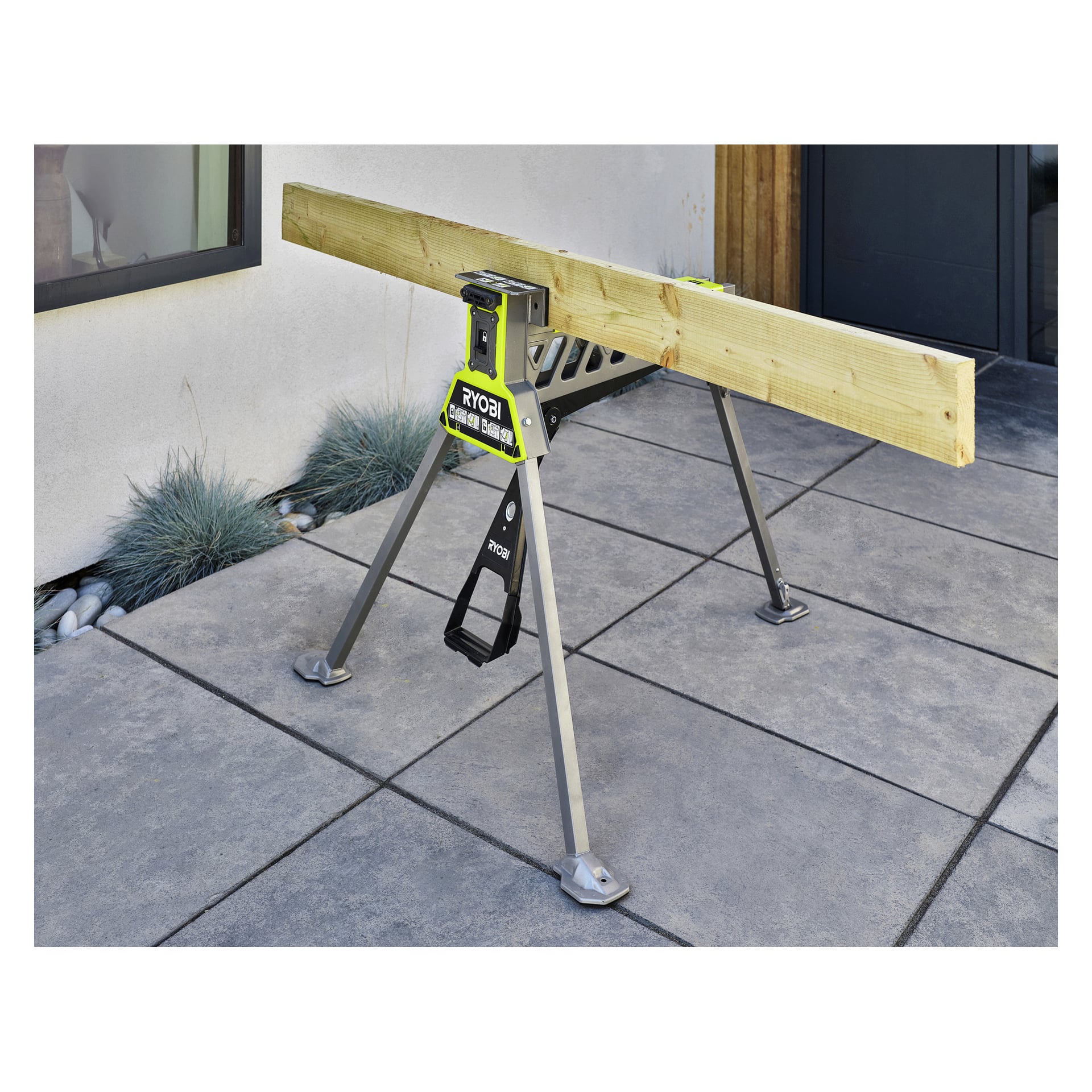 En Ryobi One+ hopfällbar arbetsbänk med klämma RSTSJ150 håller en lång träbalk på en utomhusplatt uteplats bredvid ett fönster och gräs.