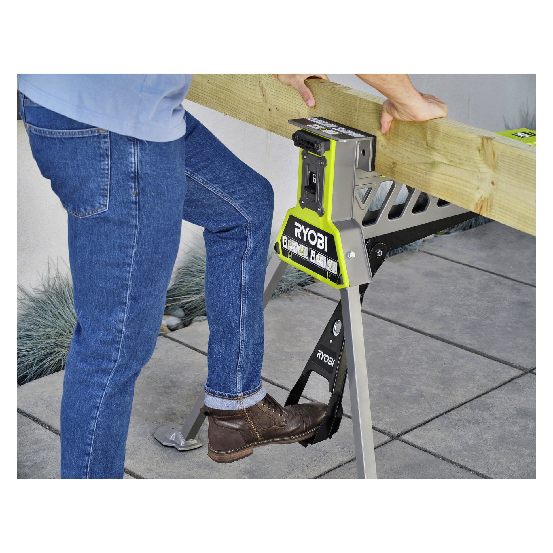 En person i blå jeans och bruna stövlar använder en Ryobi One+ Hopfällbar arbetsbänk och klämmer fast en träplanka med fot och händer.