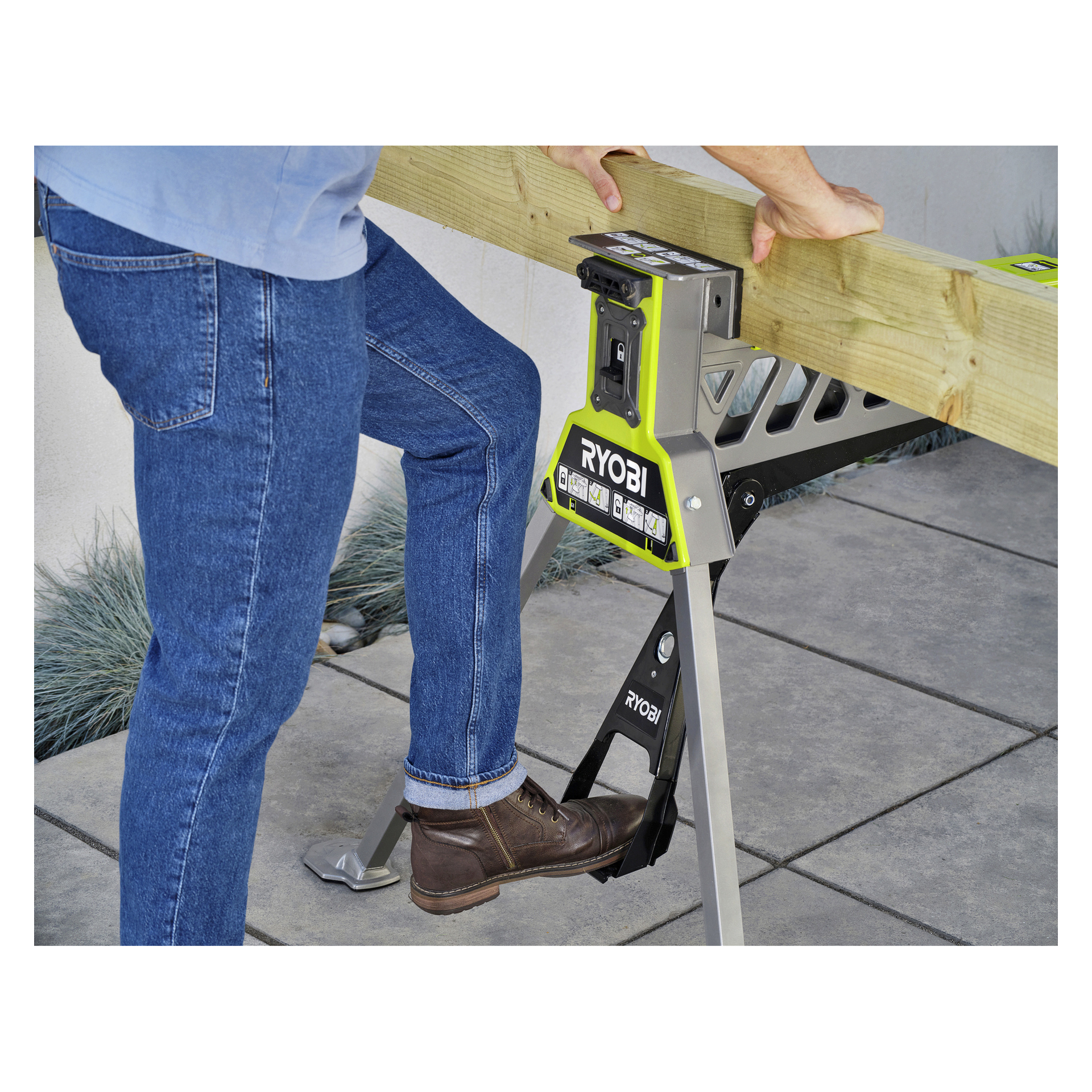 En person i blå jeans och bruna stövlar använder en Ryobi One+ Hopfällbar arbetsbänk och klämmer fast en träplanka med fot och händer.