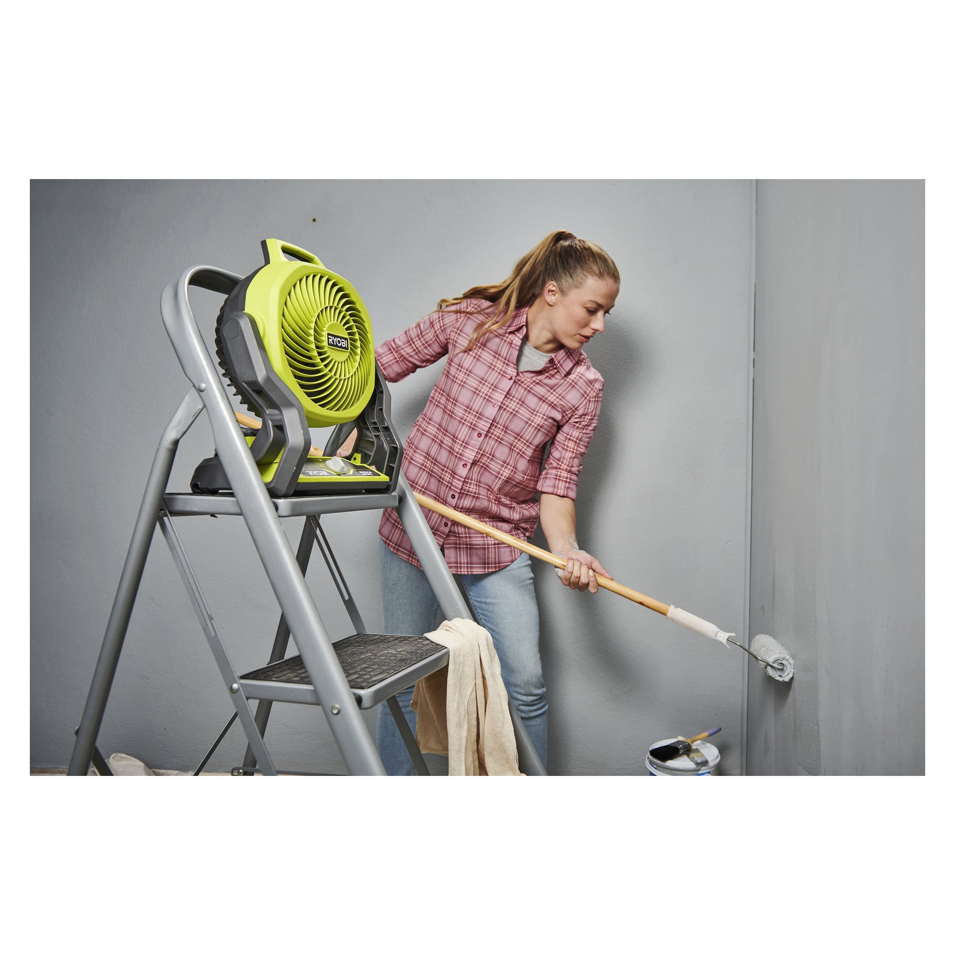 En person målar en grå vägg med en rull bredvid en stege som håller en Ryobi One+ bordsfläkt; Färgburk och pensel ligger på golvet.