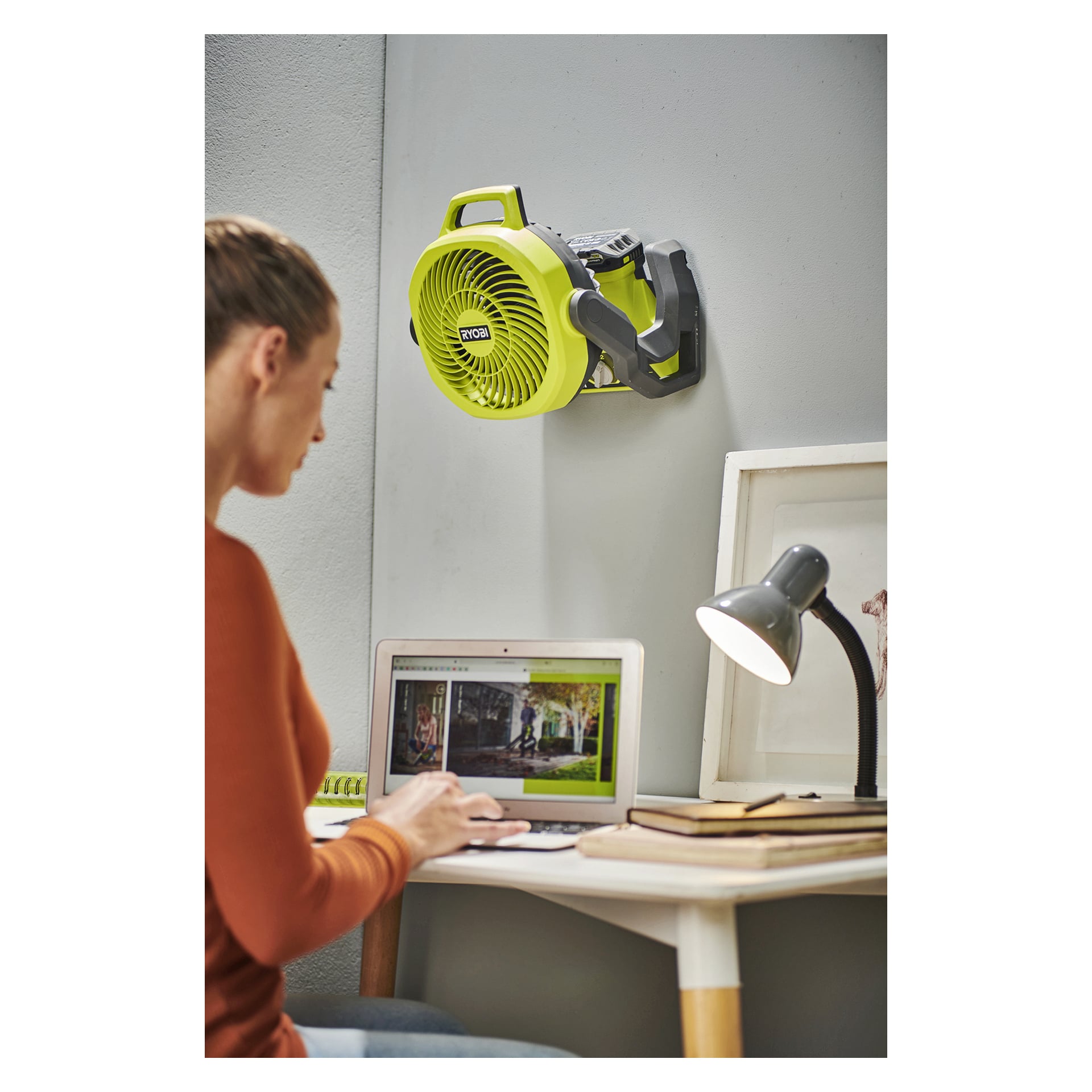 Någon arbetar vid en laptop vid ett skrivbord med en lampa, böcker och konst; ovanför är en grön Ryobi One+ RF18-0-fläkt monterad på väggen.