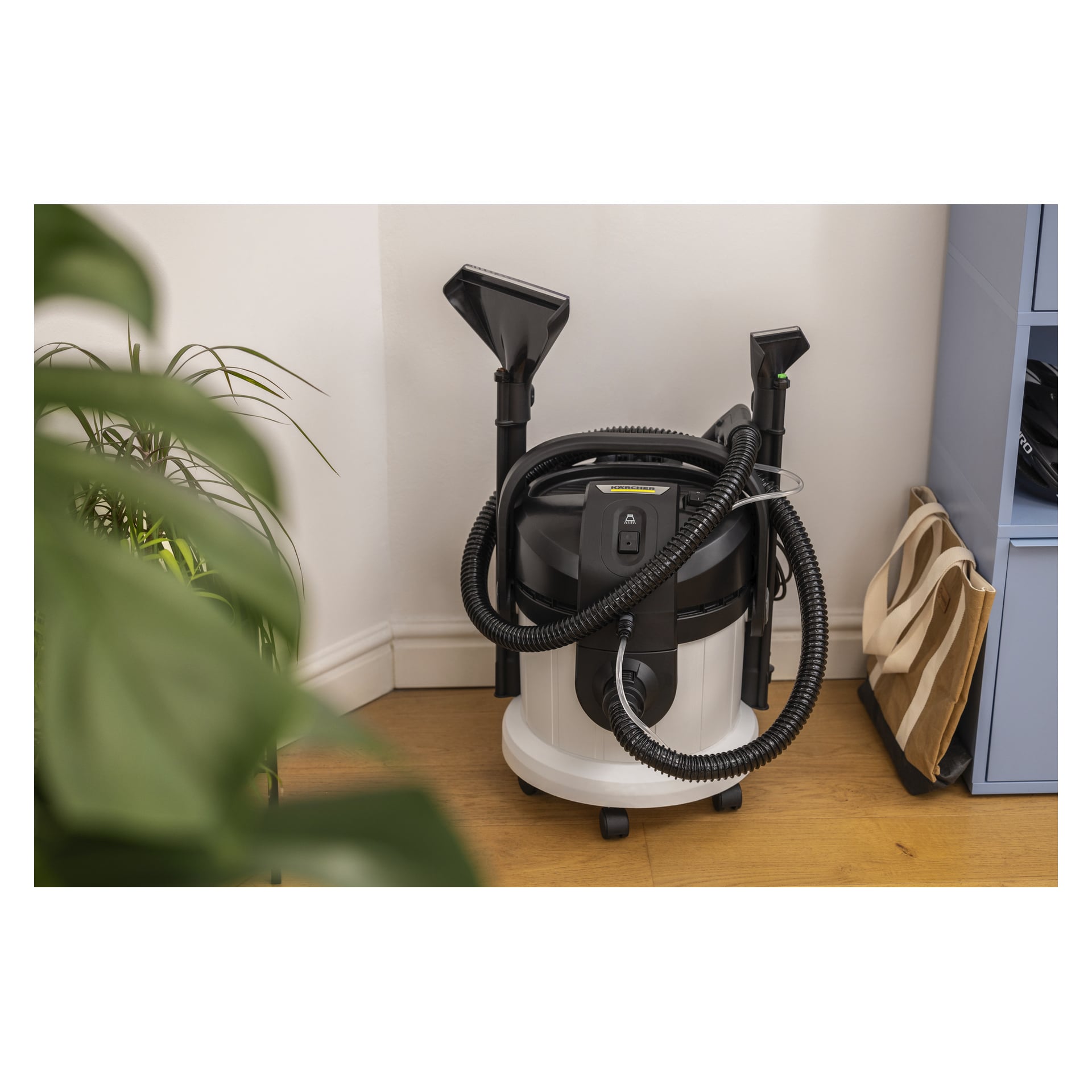 Kärcher SE 4 Plus mattrengöraren står på ett trägolv bredvid en grön växt och en hylla med påsar.