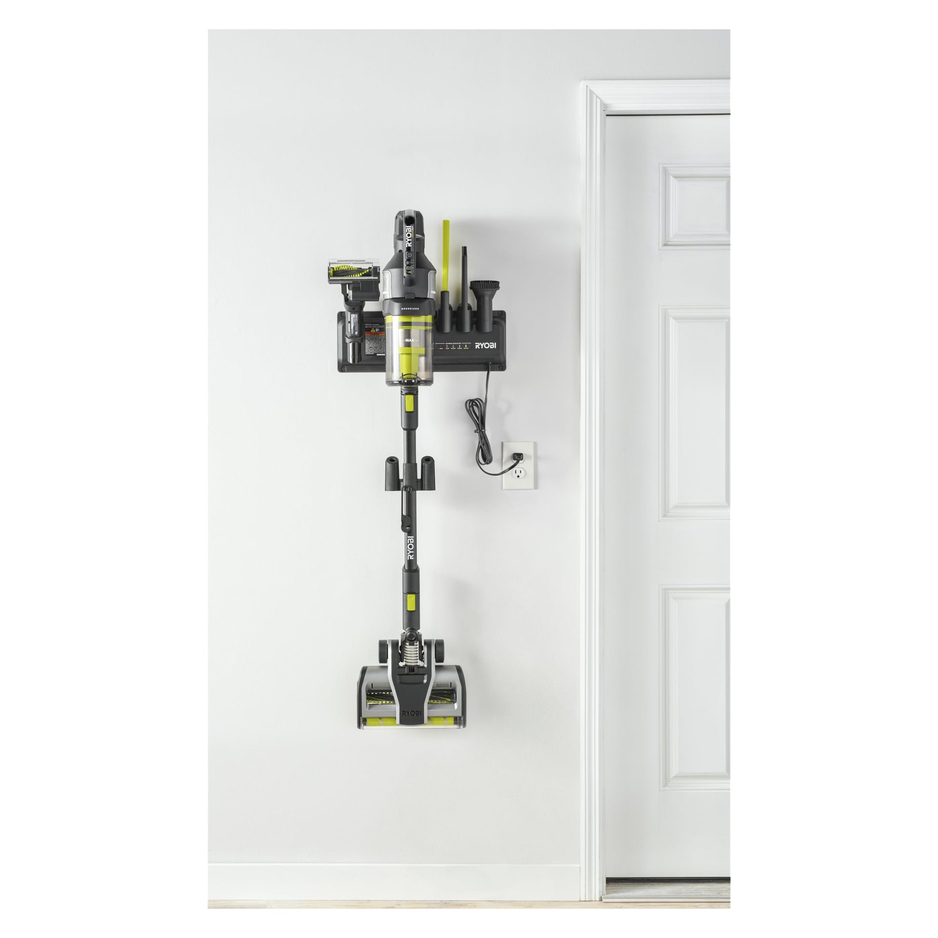 Ryobi One+ Upright dammsugare RASV18BL-1X40G på en laddstation vid en stängd dörr, strömkabeln inkopplad i ett närliggande uttag.