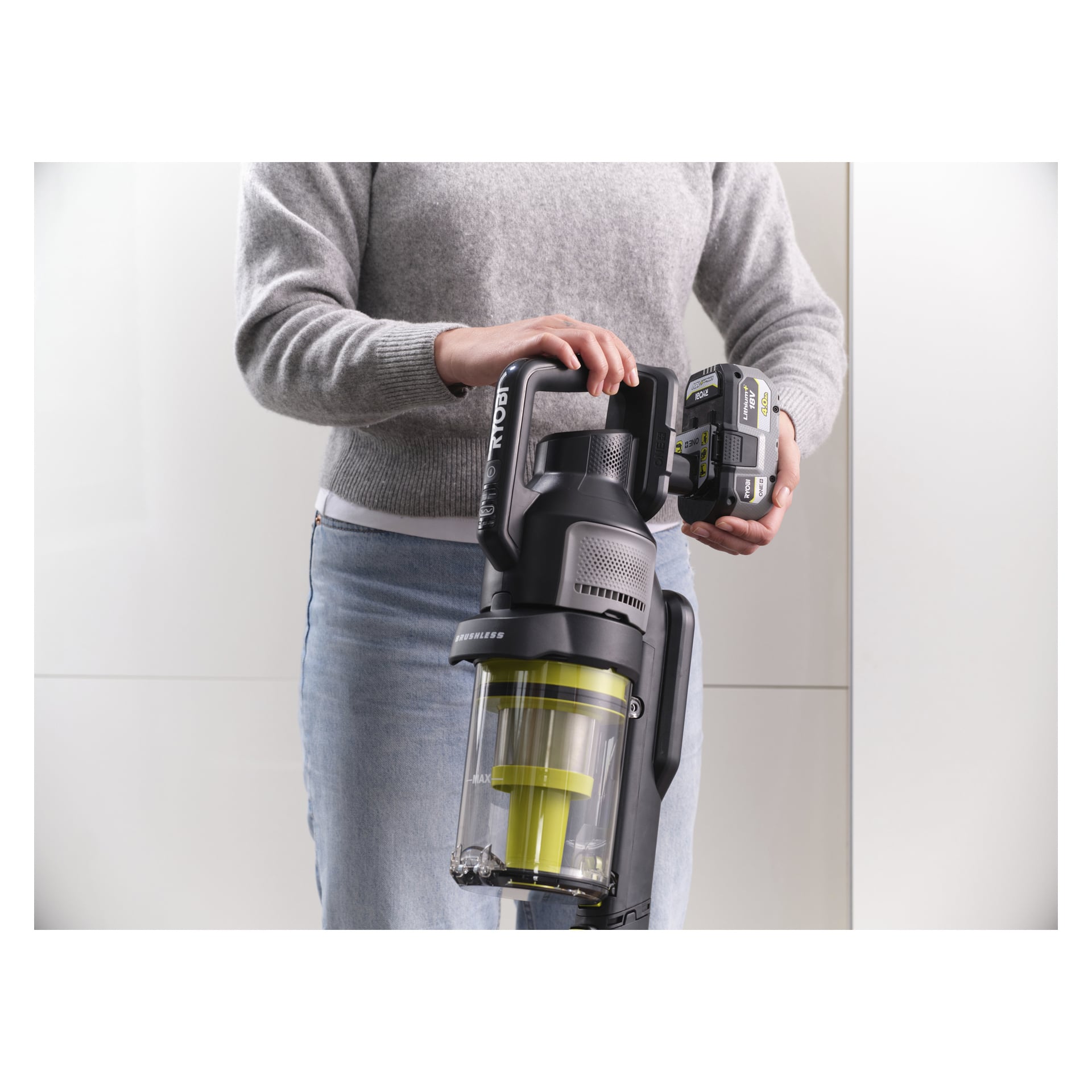 Iklädd en grå tröja och jeans håller någon en Ryobi One+ Upright dammsugare och tar ut batteripaketet med en hand.