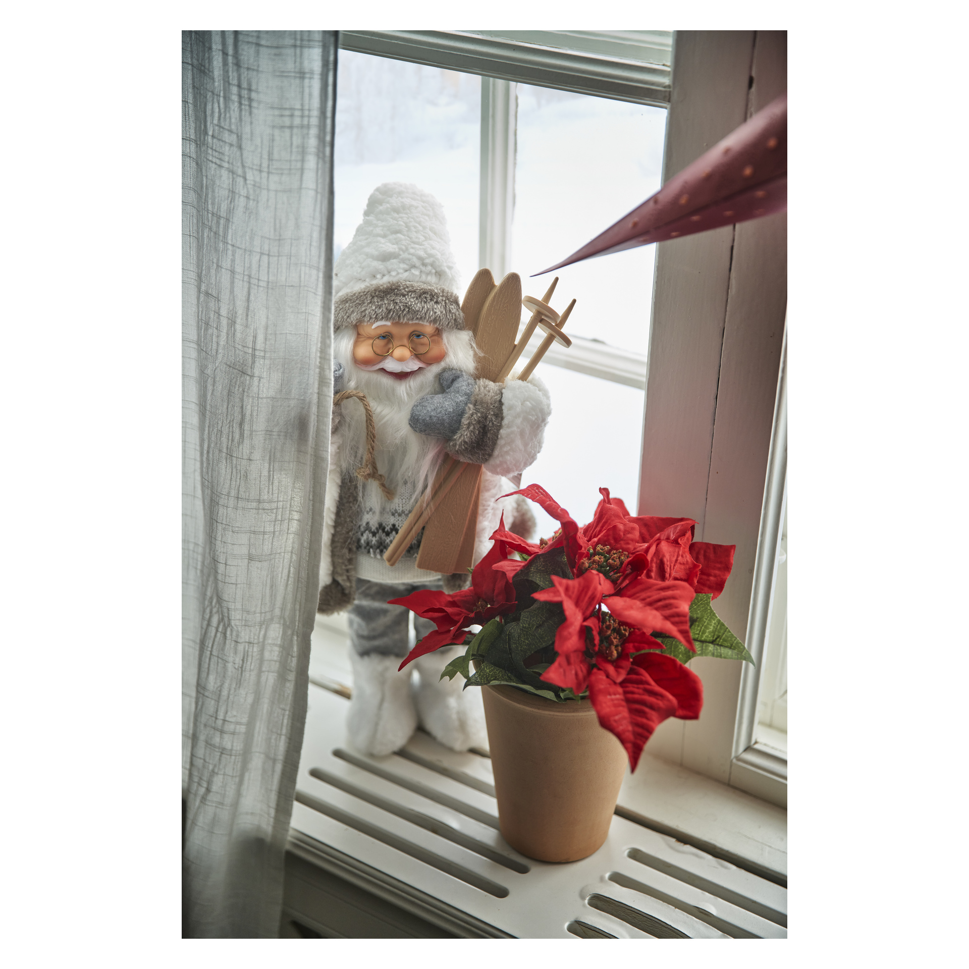 En EKVIP-tomte med skidfigur står på en fönsterbräda bredvid en julstjärna, snötäckt landskap som syns genom fönstret.
