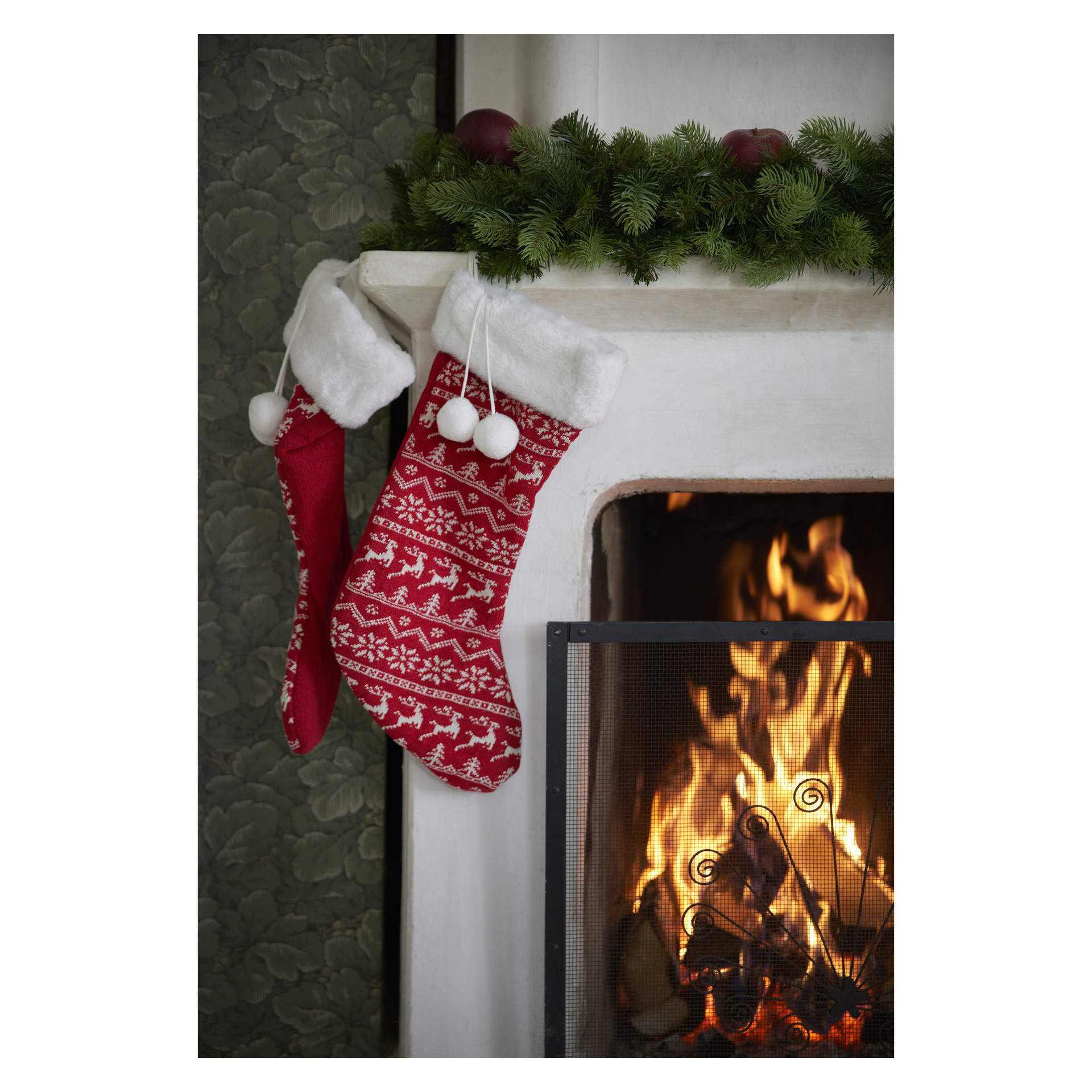 Ein Paar Menuett-Weihnachtsstrümpfe hängen an einem mit Kiefer verzierten Kaminsims über einem Kamin, der hinter einem Metallgitter leuchtet.