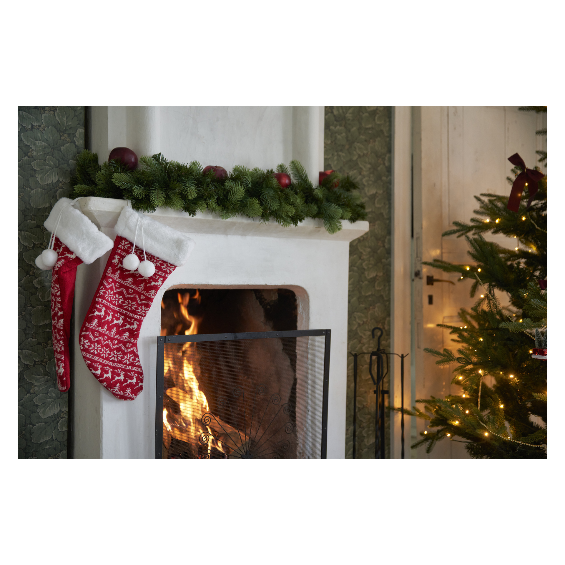 Ein Kamin mit brennendem Feuer ist mit einer grünen Girlande, roten Ornamenten, zwei rot-weißen Strümpfen und einem beleuchteten Weihnachtsbaum in der Nähe geschmückt.