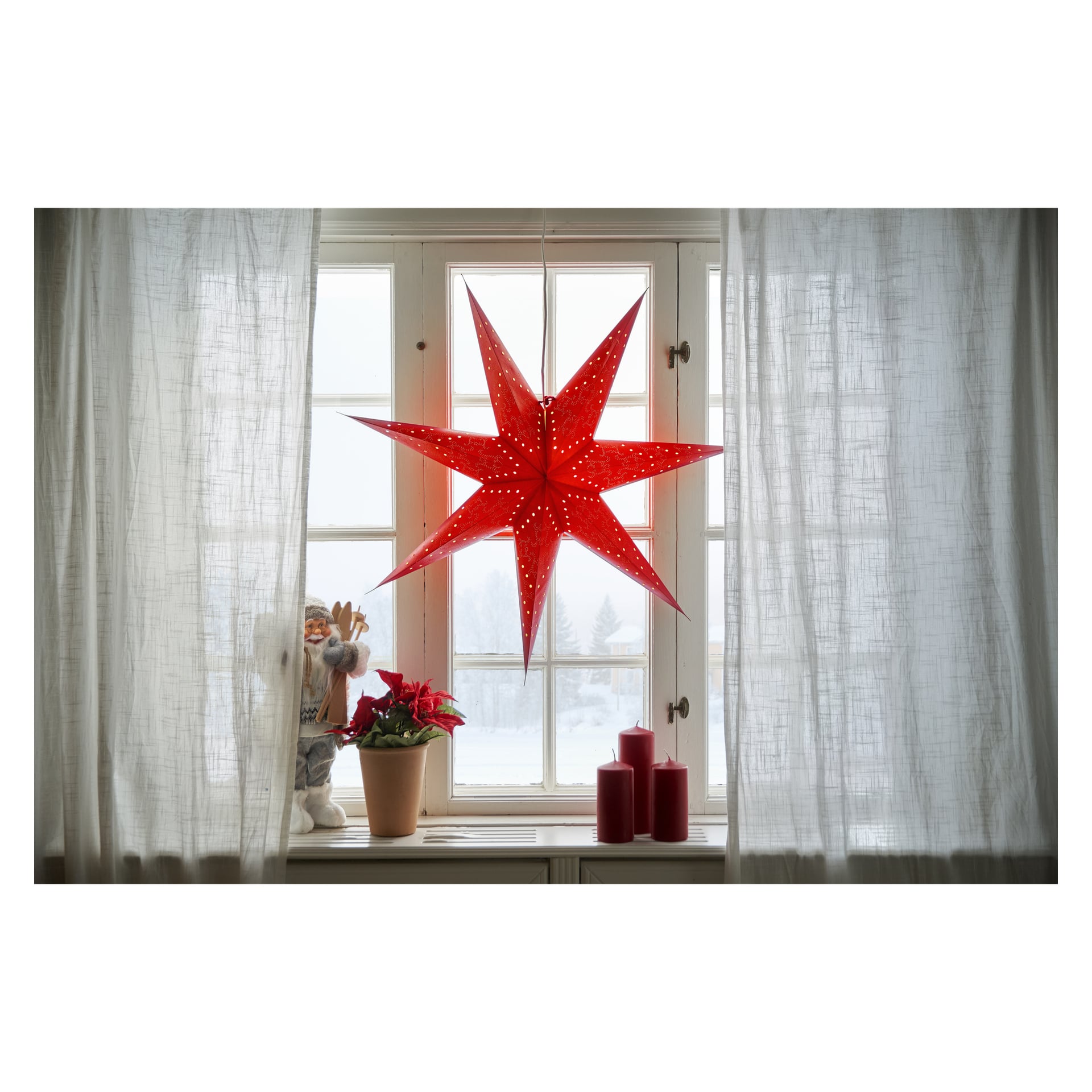 En röd adventsstjärna från EKVIP hänger framför gardinerna. På fönsterbrädan står en tomtefigur, en julstjärna i en kruka och tre röda ljus, med snö synlig utanför.