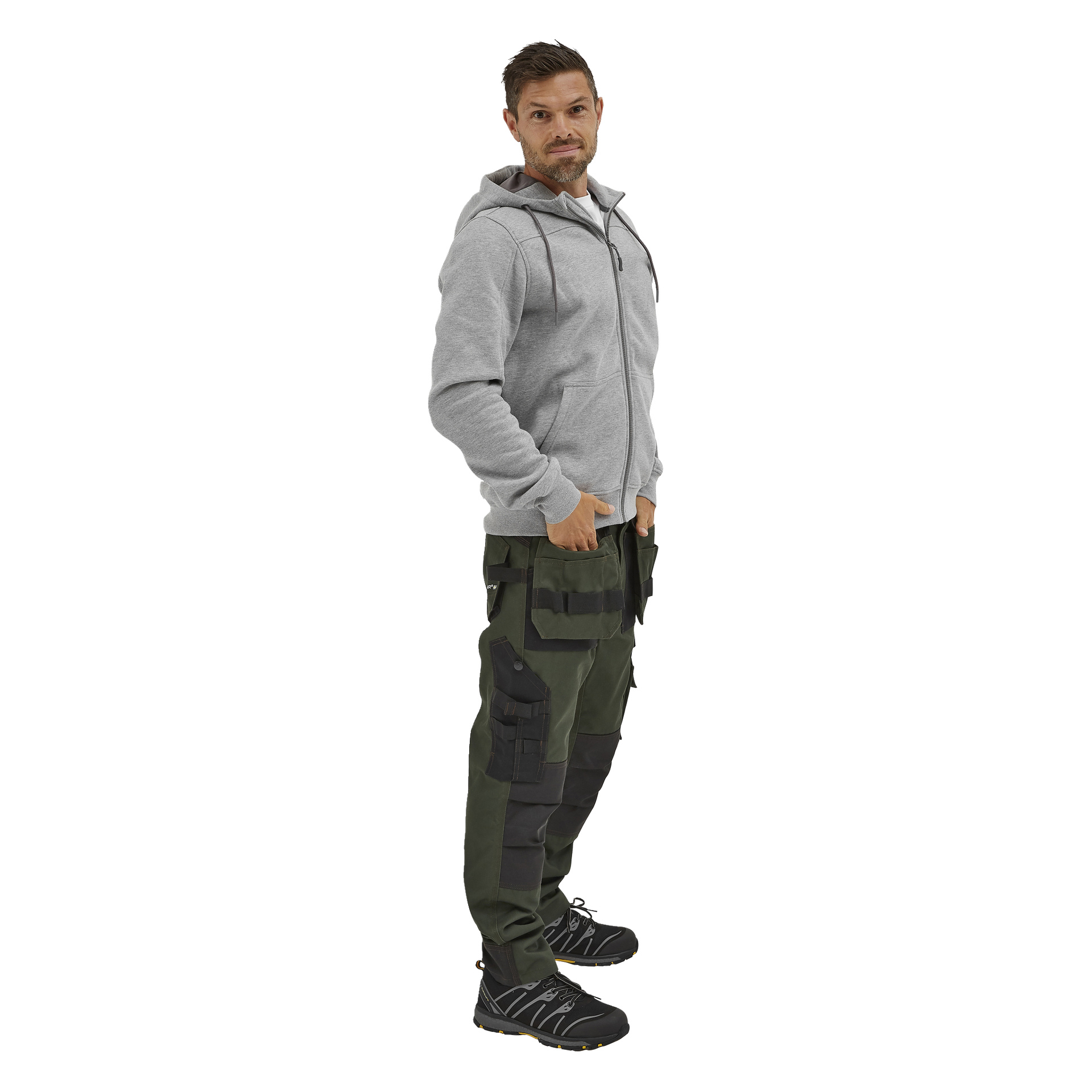 En man med kort hår och stubb står på sidan, händerna i fickorna, klädd i en ljusgrå hoodie, grön-svarta cargobyxor och svarta arbetsskor.