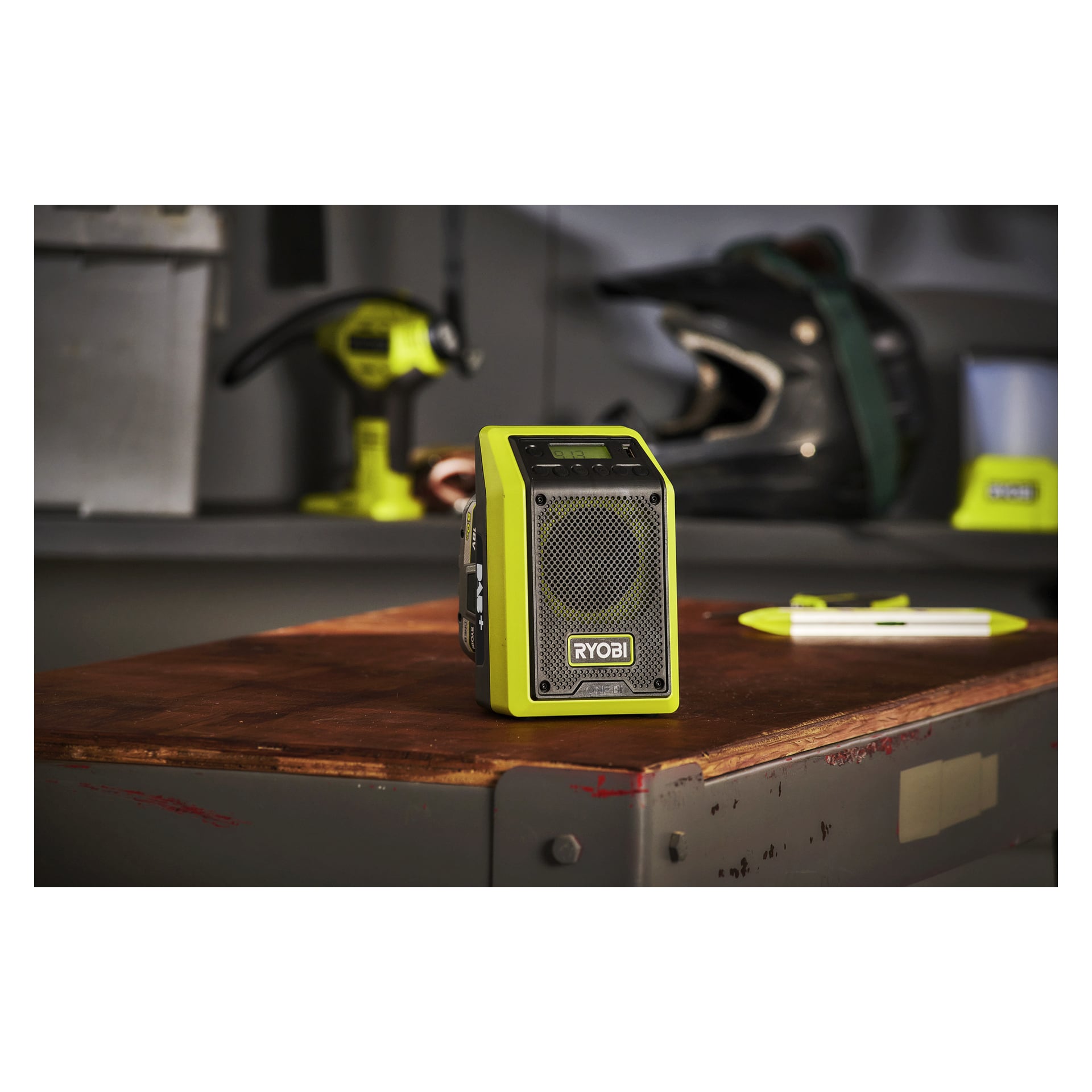 En Ryobi One+ Bluetooth-högtalare med radio RRDAB18-0 står på en träarbetsbänk, suddiga verktyg och en hjälm i verkstadens bakgrund.