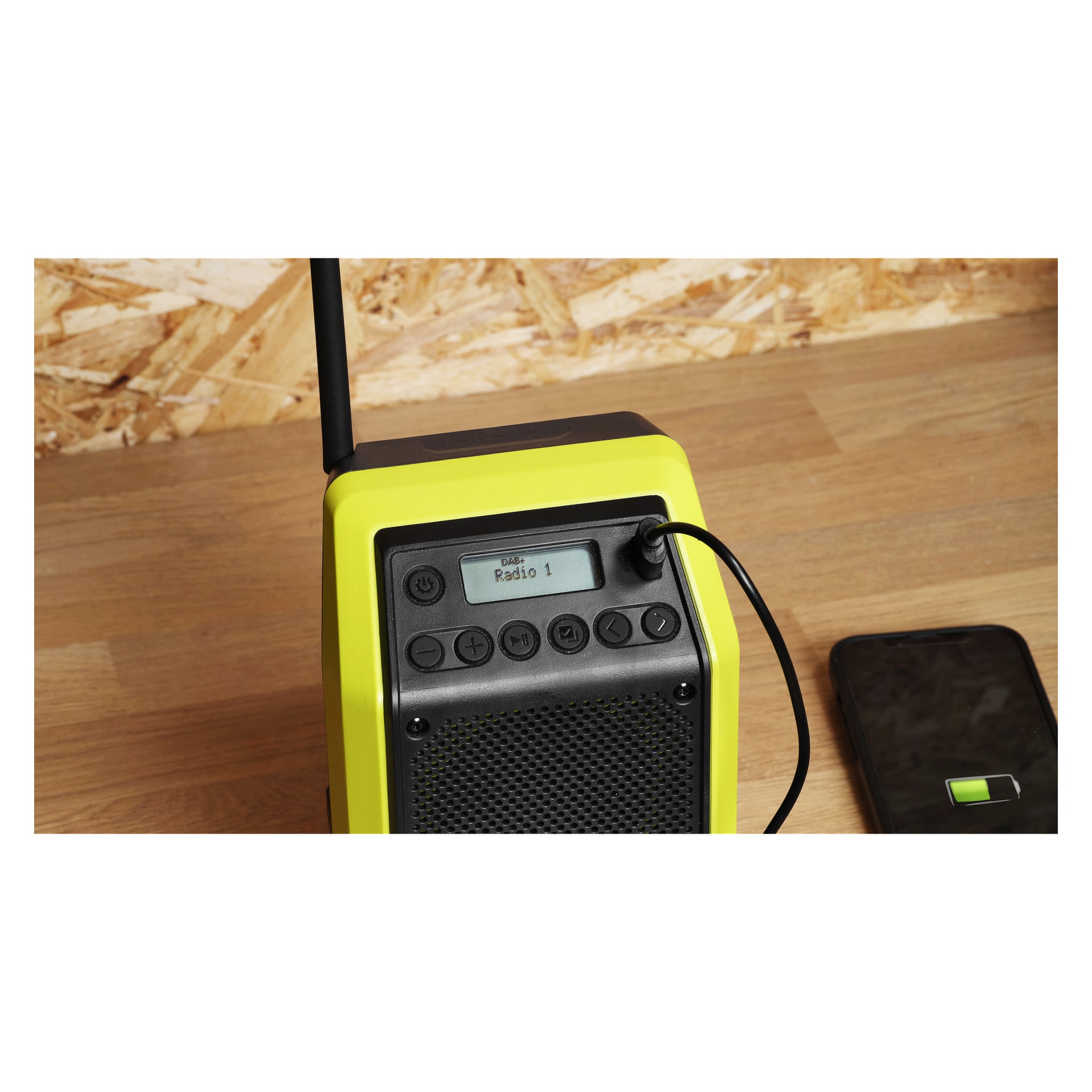 En gul och svart Ryobi One+ Bluetooth-radio står på en träyta med Radio 1 visad, kabel ansluten och telefon i närheten.