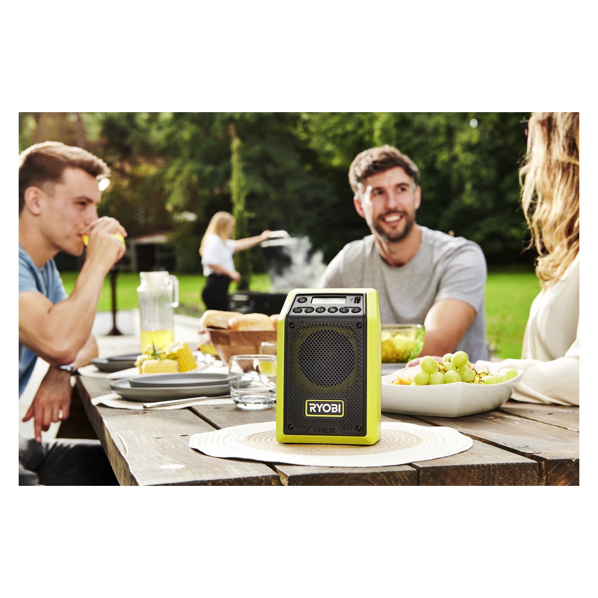 En Ryobi One+ Bluetooth-högtalare med radio RRDAB18-0 står på ett utomhusbord i trä med mat och dryck, folk som pratar i närheten och någon som grillar på gräsmattan.