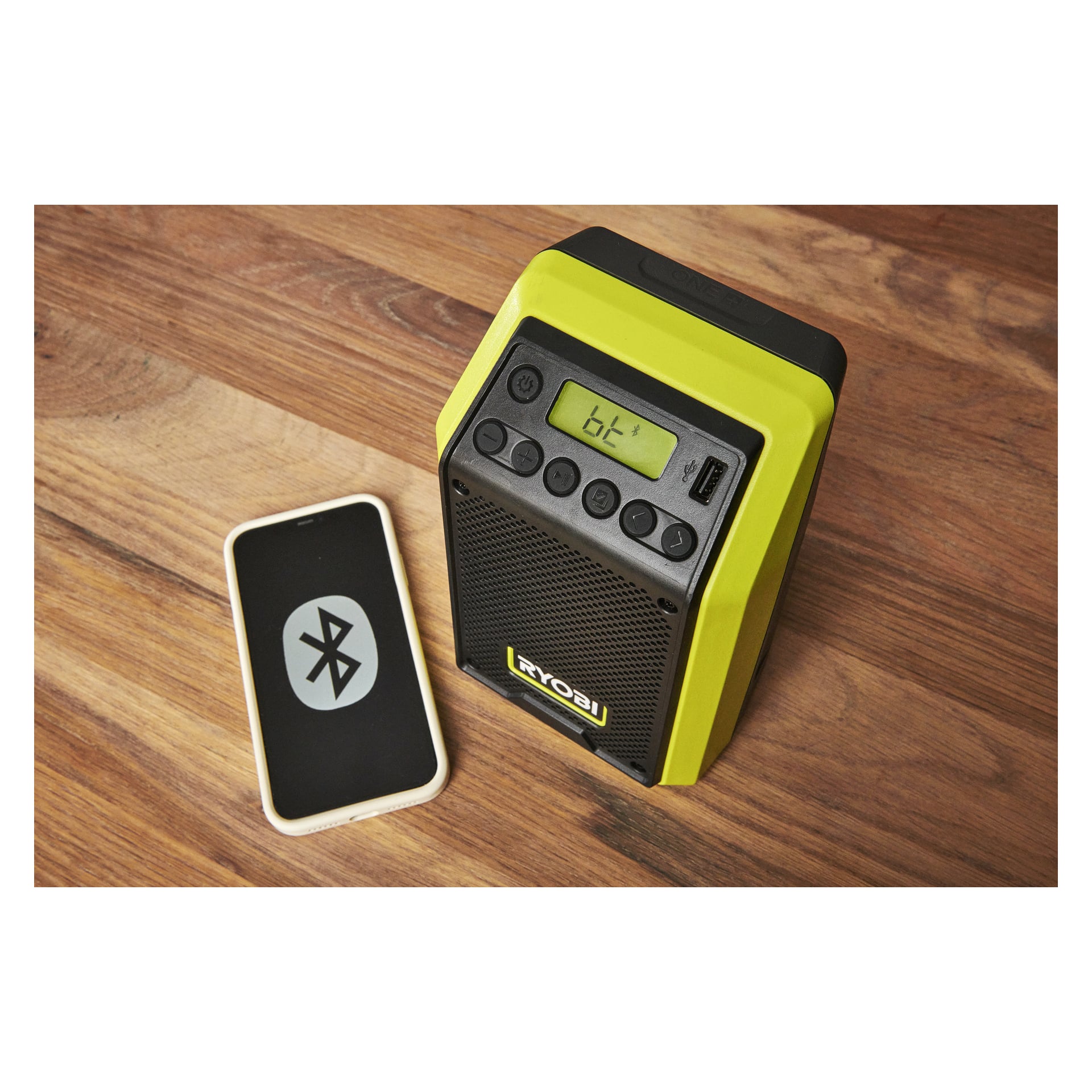 En gul och svart Ryobi One+ Bluetooth-högtalare med radio RRDAB18-0 vilar på trä bredvid en smartphone som visar en Bluetooth-ikon.