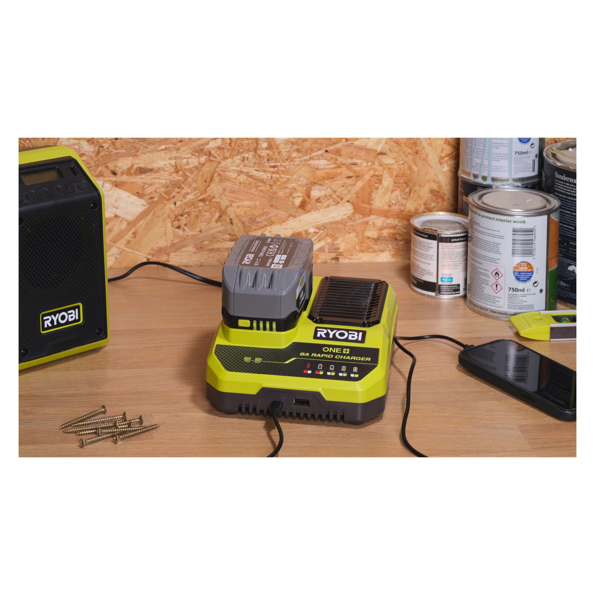 En Ryobi One+ Quick laddare RC18180 att ladda ett batteri på en träarbetsbänk med närliggande skruvar, telefon, färgburkar och högtalare.