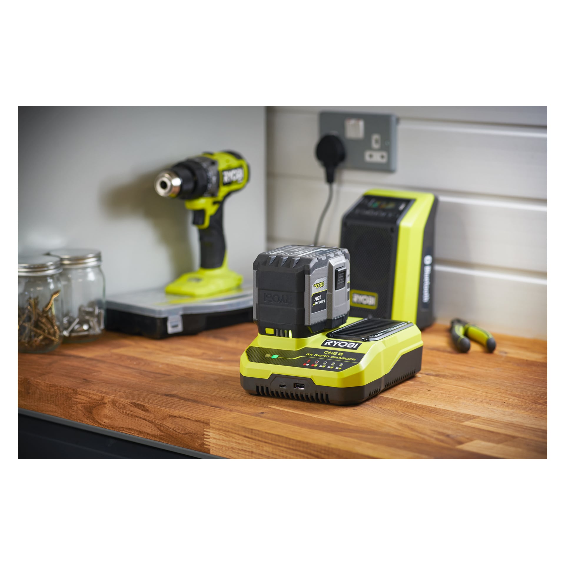 En Ryobi One+ Quick-laddare laddar RC18180 ett batteri på en träarbetsbänk med borrmaskin, högtalare, skruvburkar och tång i närheten.