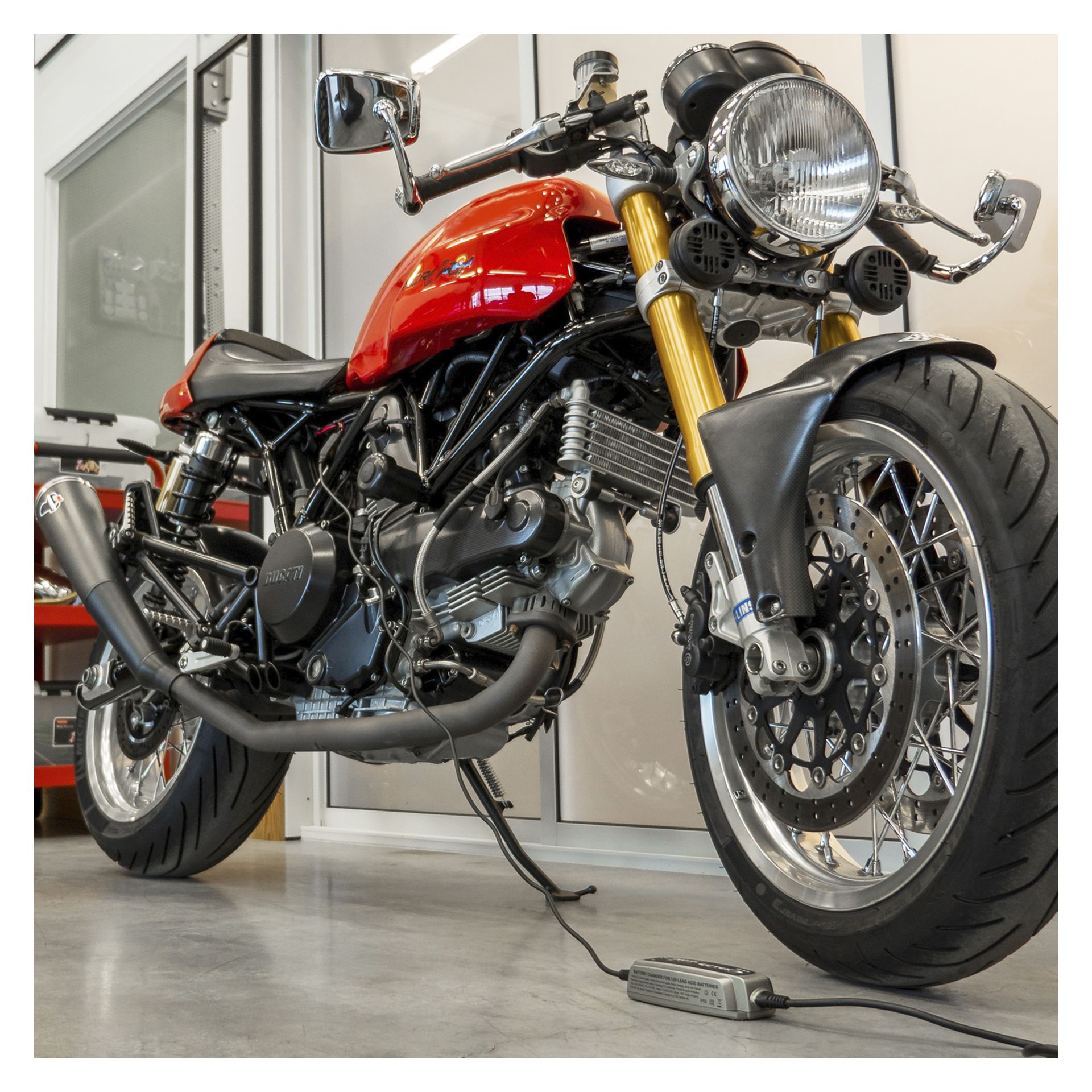 Czerwono-motocykl ze złotymi przednimi widelcami stoi w środku na szarej podłodze, podłączony do ładowarki baterii CTEK XS 0.8.