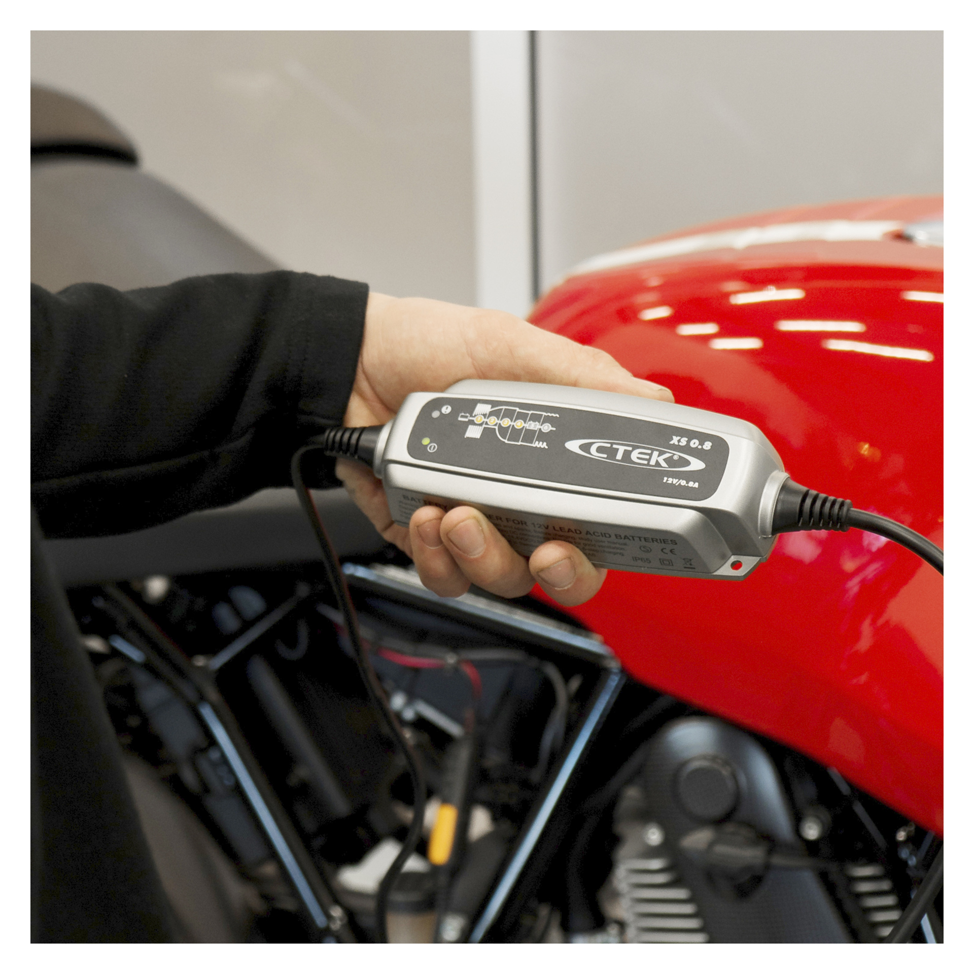 Ręka trzyma ładowarkę baterii CTEK XS 0.8 obok motocykla, z widocznym silnikiem i zbiornikiem paliwa za nimi.