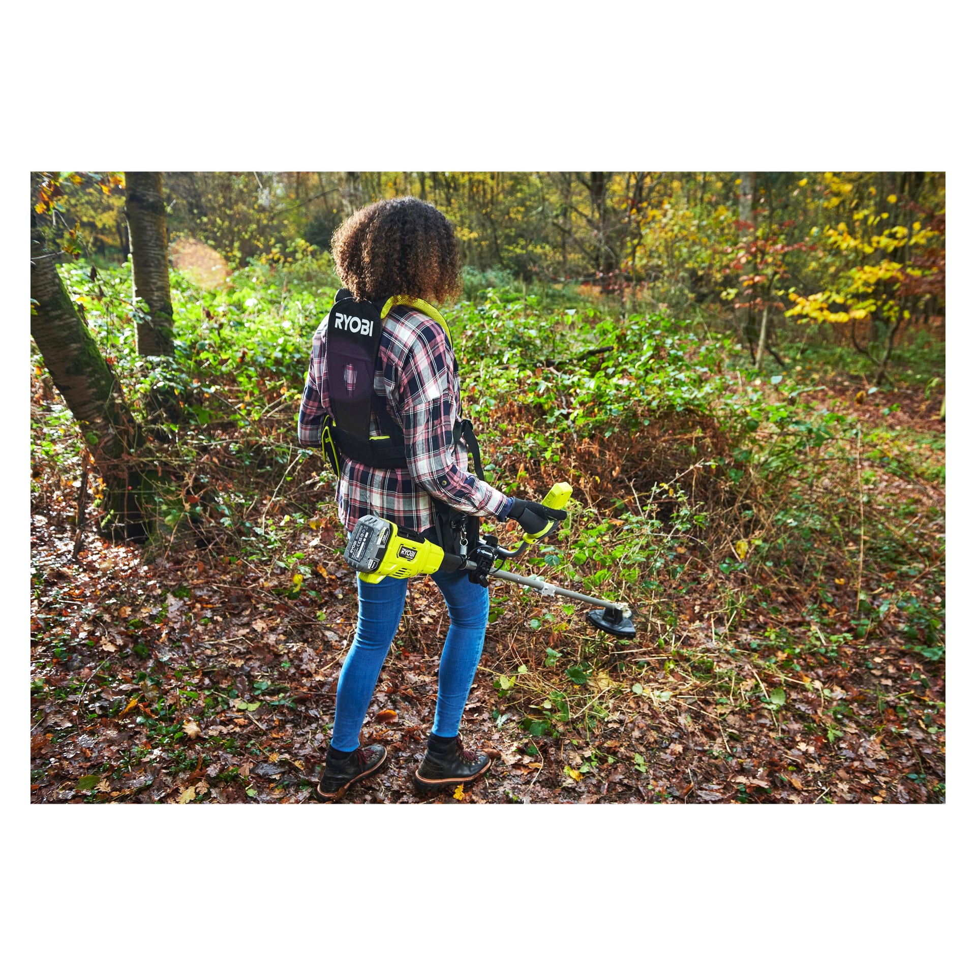 Iført rutete skjorte og jeans klipper man tett undervegetasjon blant falne blader med en Ryobi One+ HP børsteklipper i skogen.