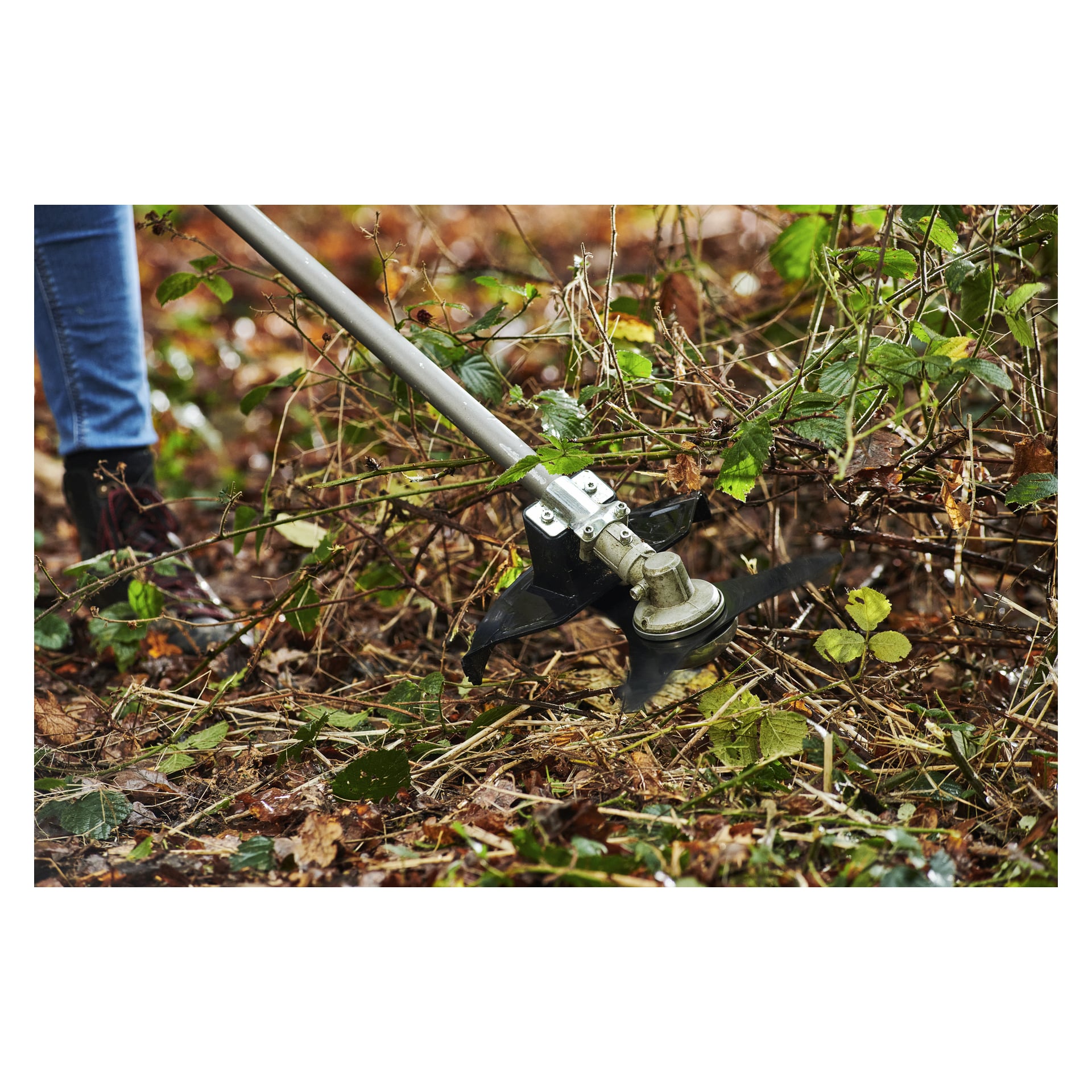 En person bruker en Ryobi One+ HP batteribørsteskjærer med metallblad for å kutte tett kratt; bare beinet og støvelen er synlige.