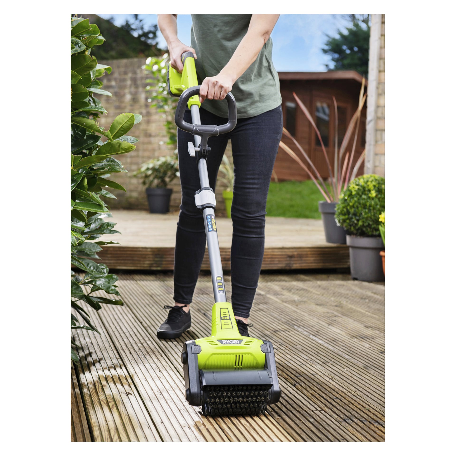 Iklädd en grön skjorta och svarta byxor rengör en person trädäck med en Ryobi One+ Patio Cleaner, trädgård och skjul bakom.