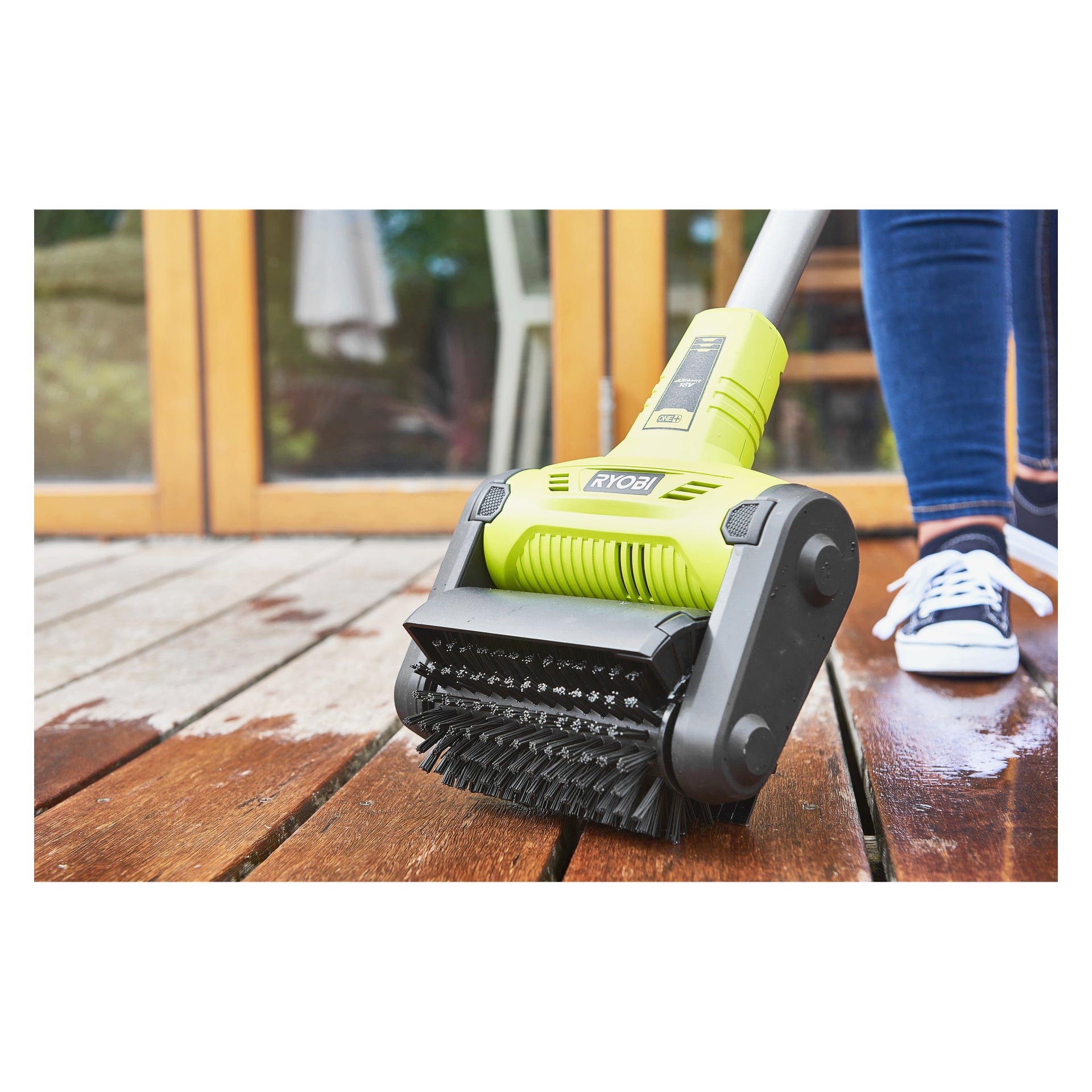 En person i jeans och sneakers rengör en träaltan nära glasdörrar med en Ryobi One+ uteplatsrengörare RY18PCB-140W.