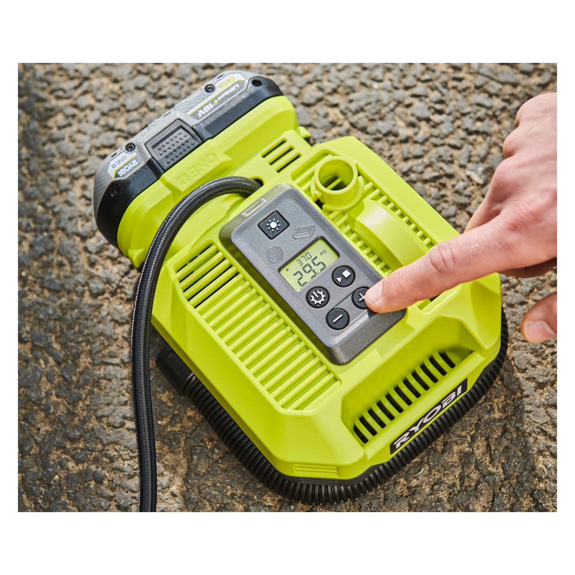 En person använder en Ryobi One+ batteridriven luftkompressor på texturerad asfalt, med en digital display som visar 24,9 PSI.