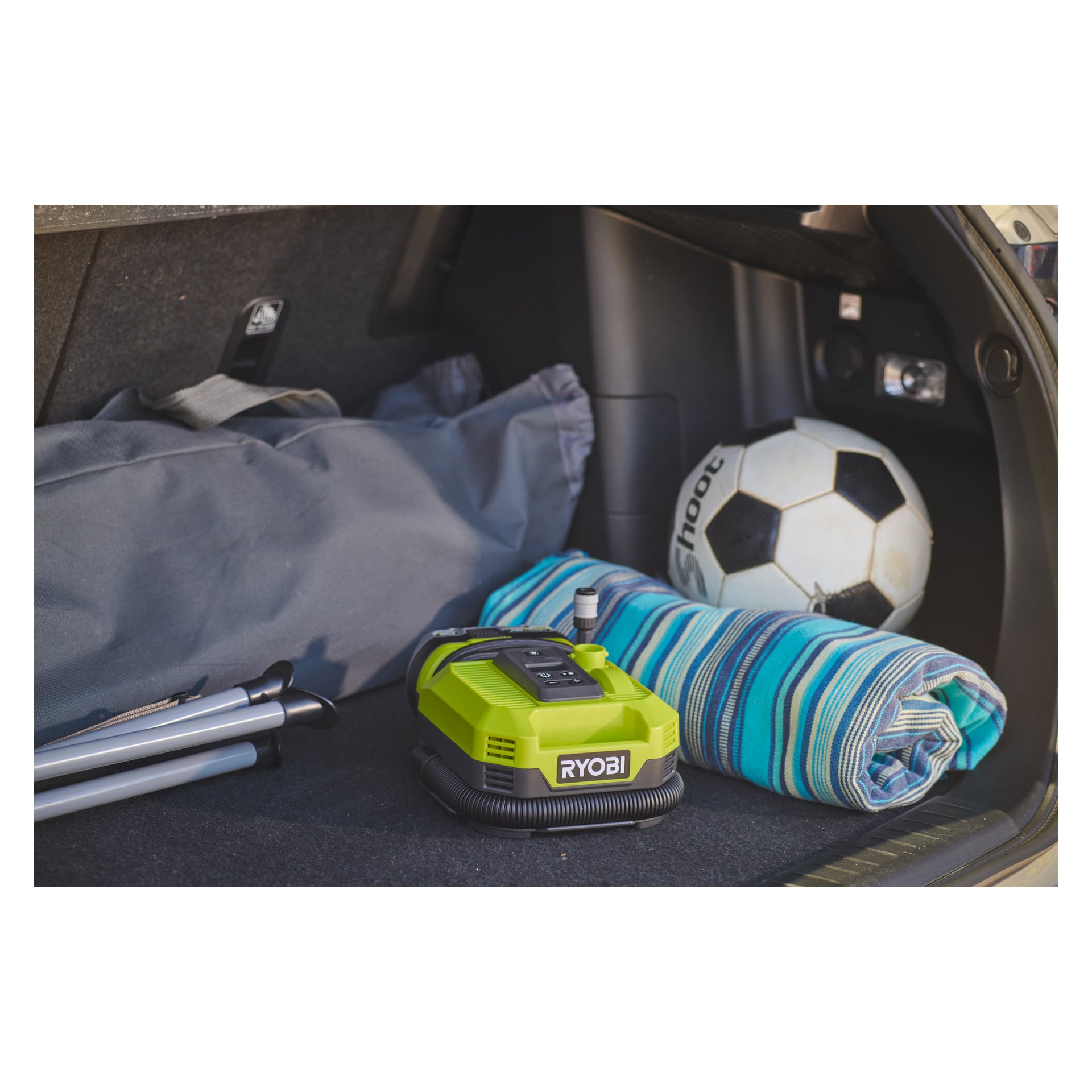 En bilbagageutrymme rymmer ett vikt stativ, grå väska, randig blå filt, fotboll och en Ryobi One+ Battery multifunktionskompressor.