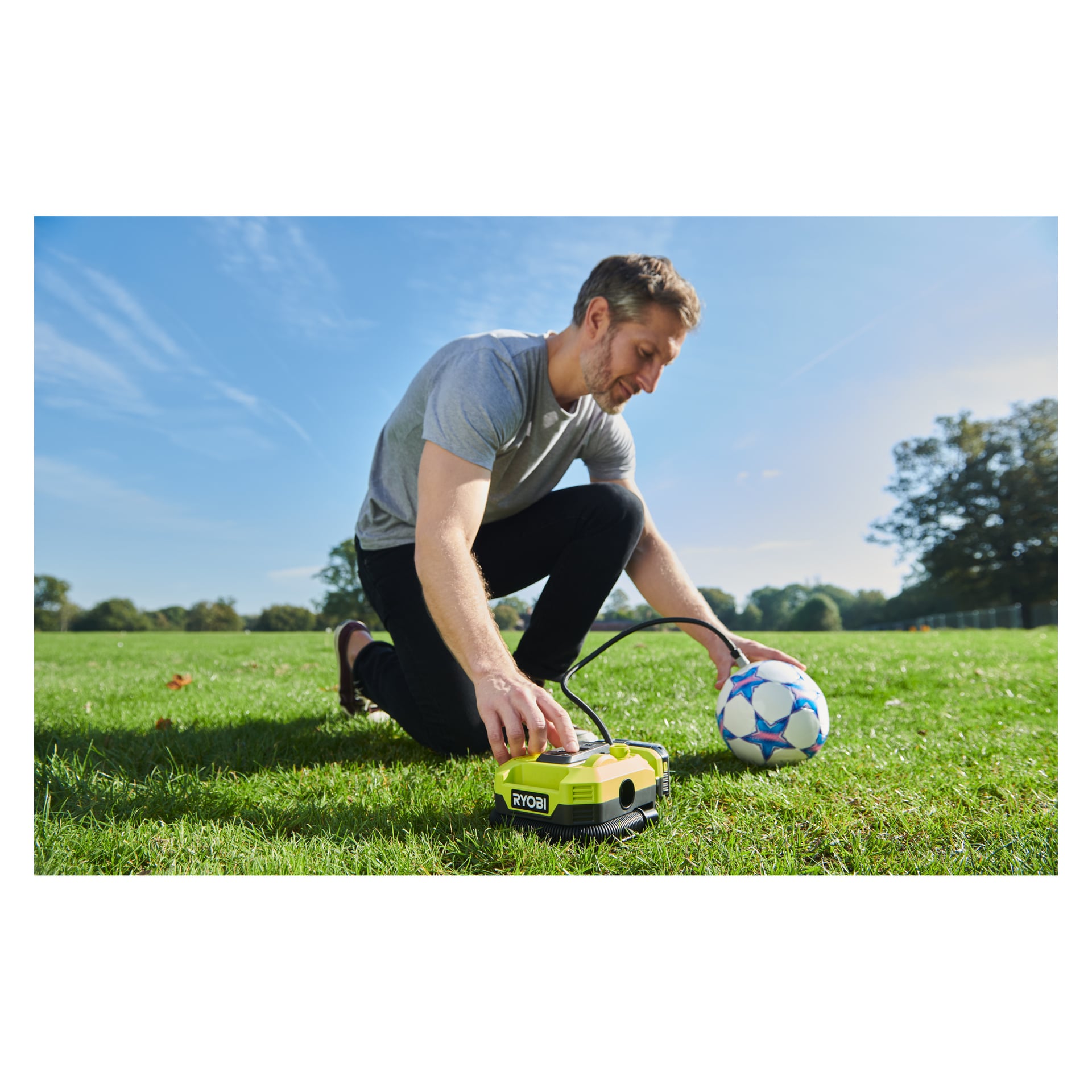 En man knäböjer på parkgräset och blåser upp en fotboll med en Ryobi One+ luftkompressor under klar himmel och träd.