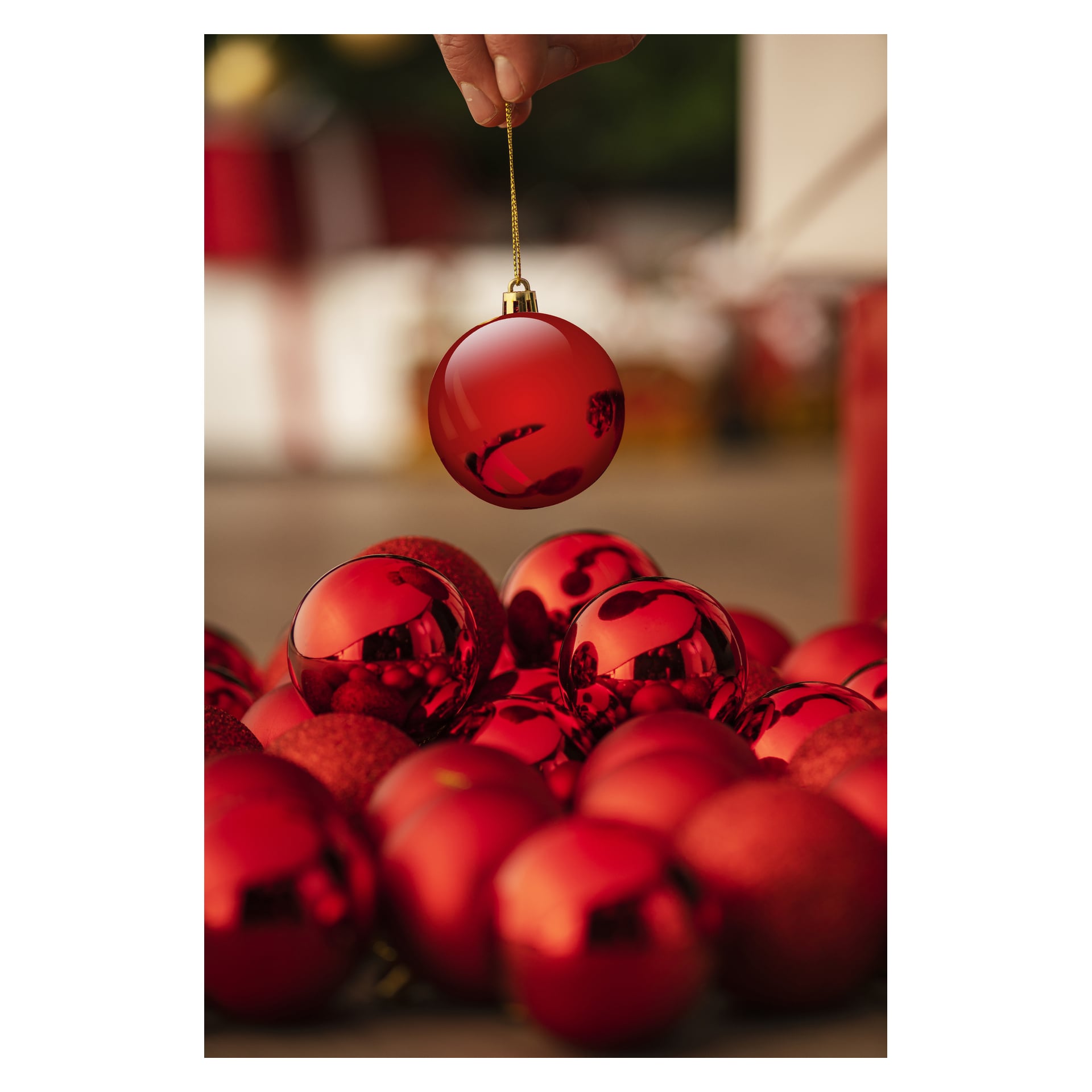 En hand håller en prydnad märkt "EKVIP Christmas tree balls 20 pack" i sitt snöre över en grupp röda prydnader på en yta. Scenen är festlig, med huvudfokus på prydnaderna och en mjukt suddig bakgrund.