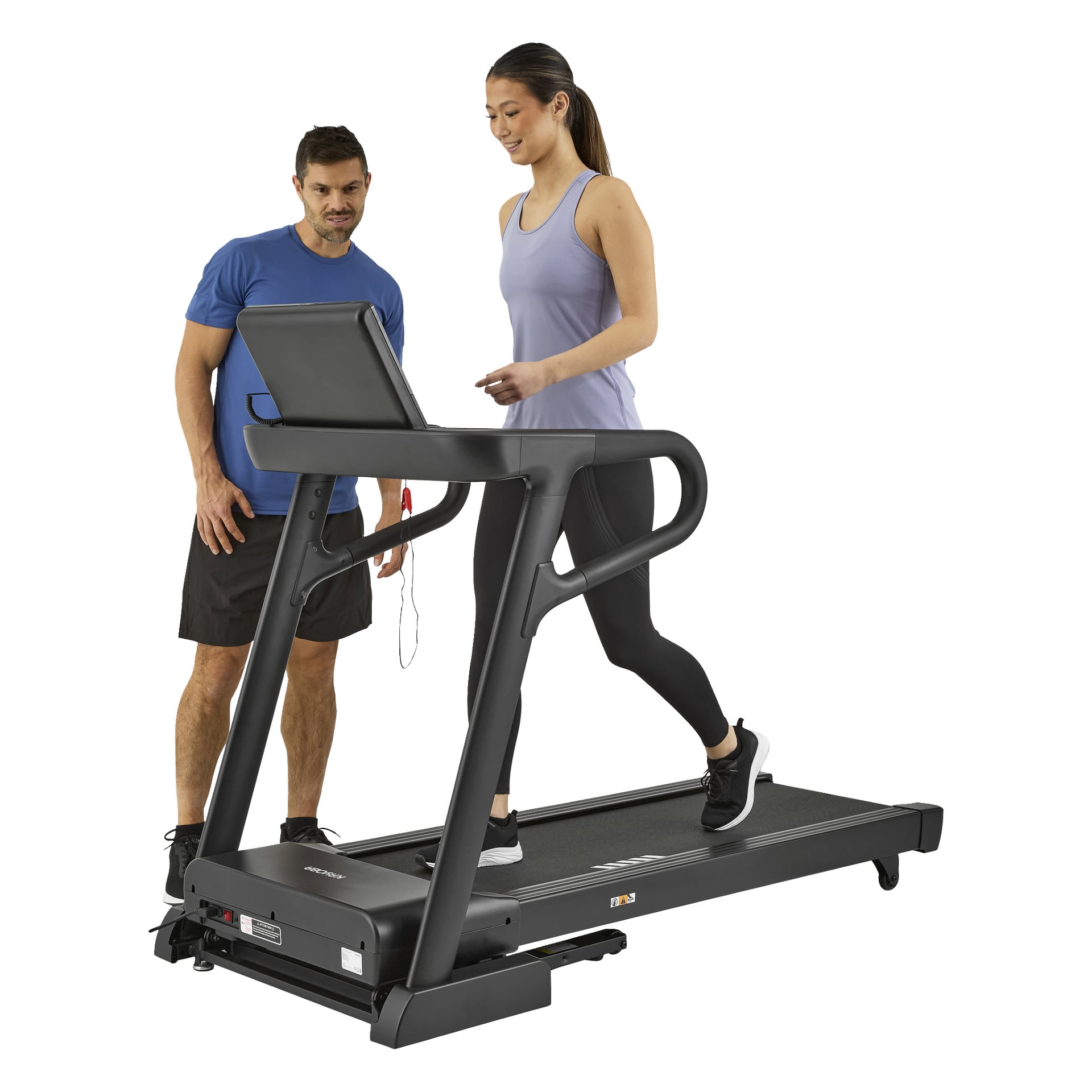 En kvinna tränar på ett Kayoba-löpband med lutning, medan en man i sportkläder står bredvid henne och kollar på utställningen i ett gym.