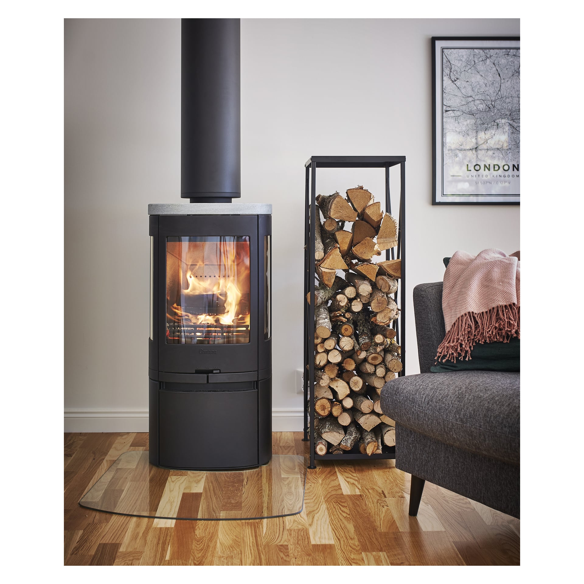 En elegant svart vedovn lyser ved siden av vedfyllte Anslut Firewood-lager. En grå sofa med rosa pledd og et London-kart gir koselighet.