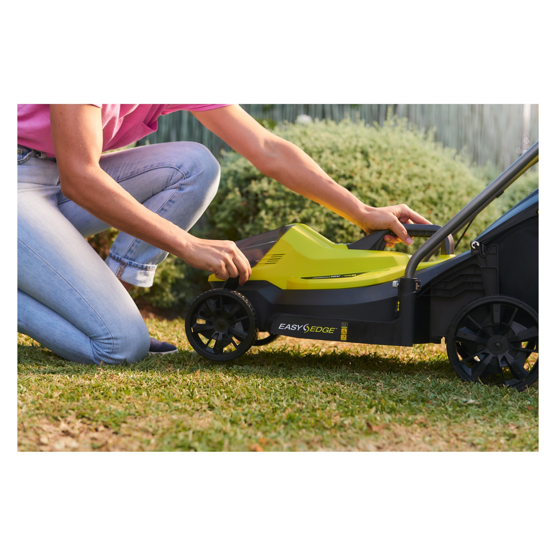 En person iført rosa skjorte og jeans setter sammen en gul og svart "Ryobi One+ batteridrevet gressklipper OLM1833B-0" på gresset, omgitt av grønt og et gjerde.