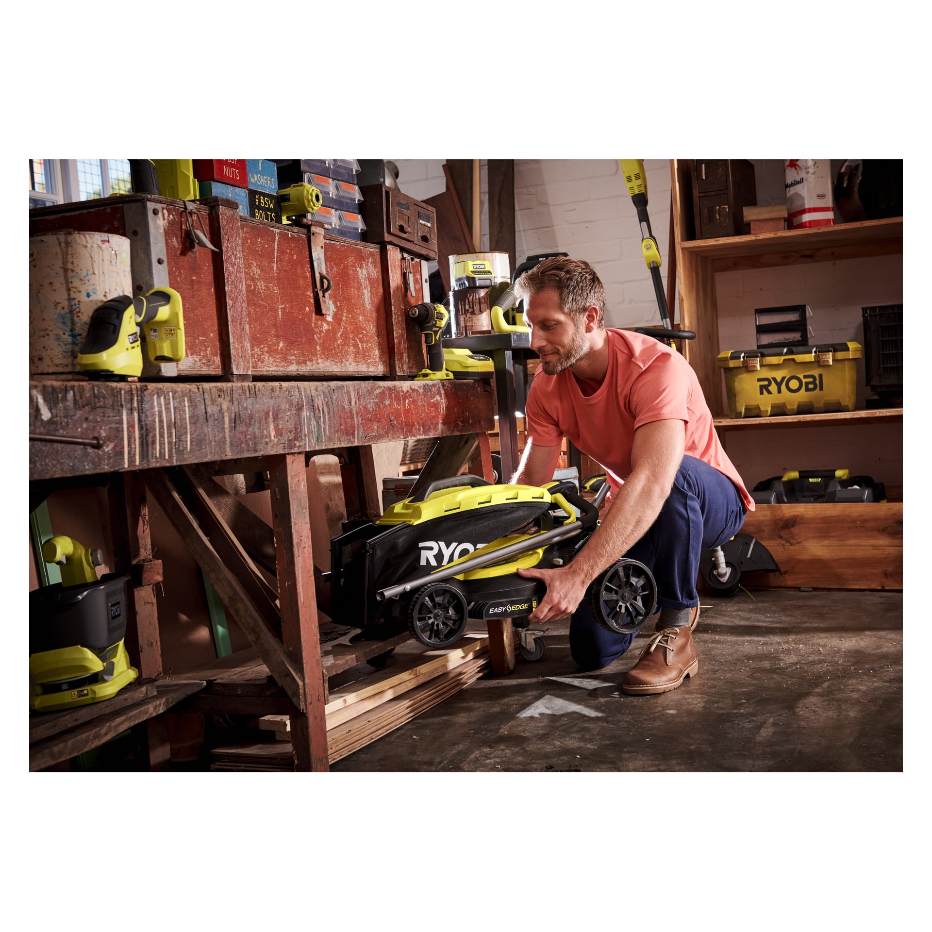 En mann kneler i et verksted og skyver en sammenbrettet «Batteridrevet gressklipper OLM1833B-0» under en arbeidsbenk i tre. Ryobi One+ verktøy og verktøykasser er plassert på hyller og benken rundt ham.