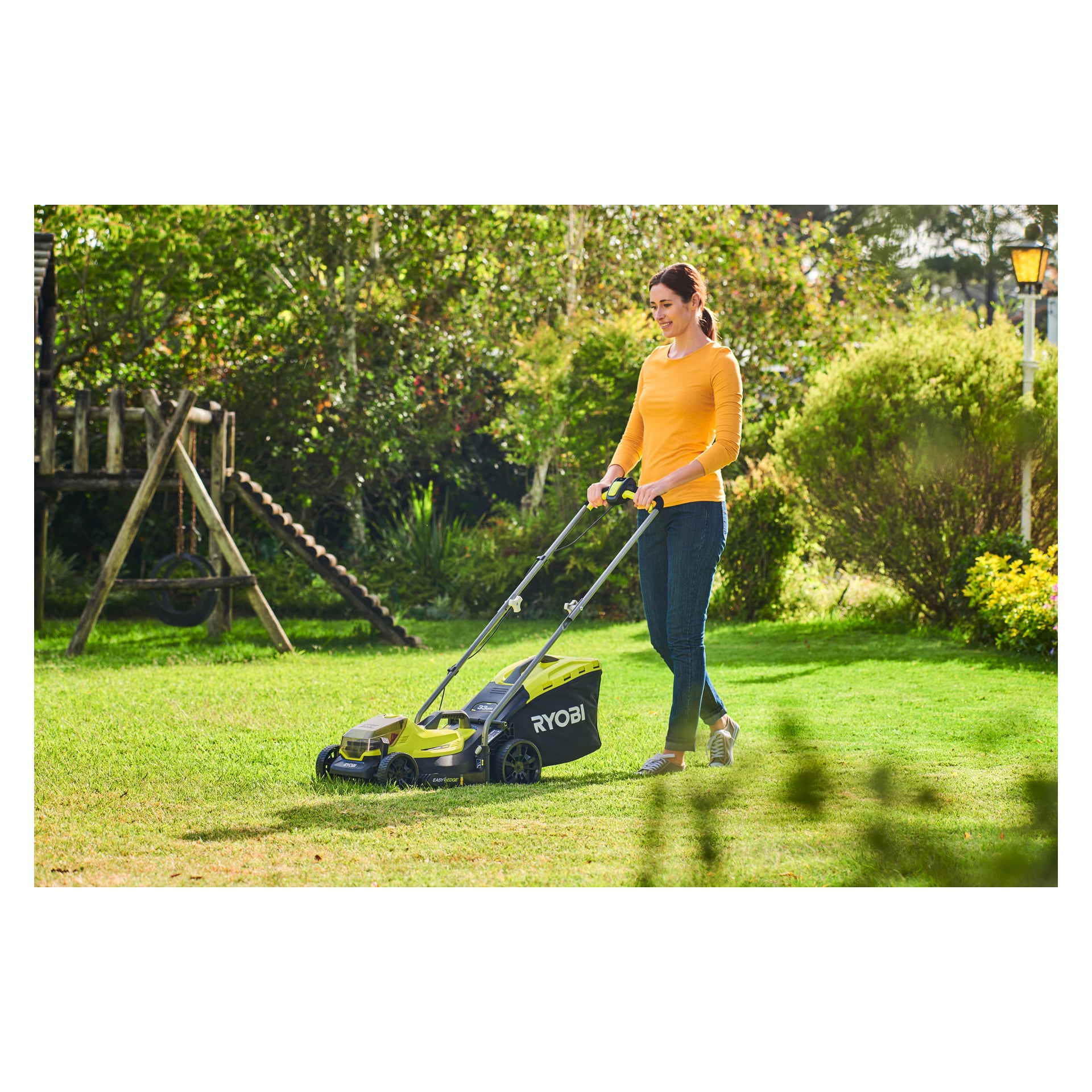 En person iført en gul langermet skjorte og jeans skyver en Ryobi One+ batteridrevet gressklipper OLM1833B-0 over en gresskledd hage med trær og busker. Solen skinner.