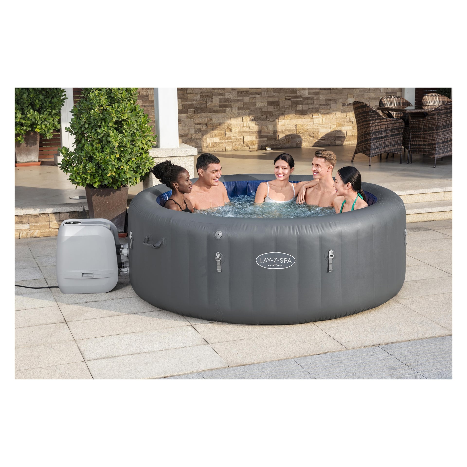 Fem personer slapper av i et rundt, grått LAY-Z-SPA Santorini Max Hold spa på en steinterrasse, omgitt av utemøbler og potteplanter.