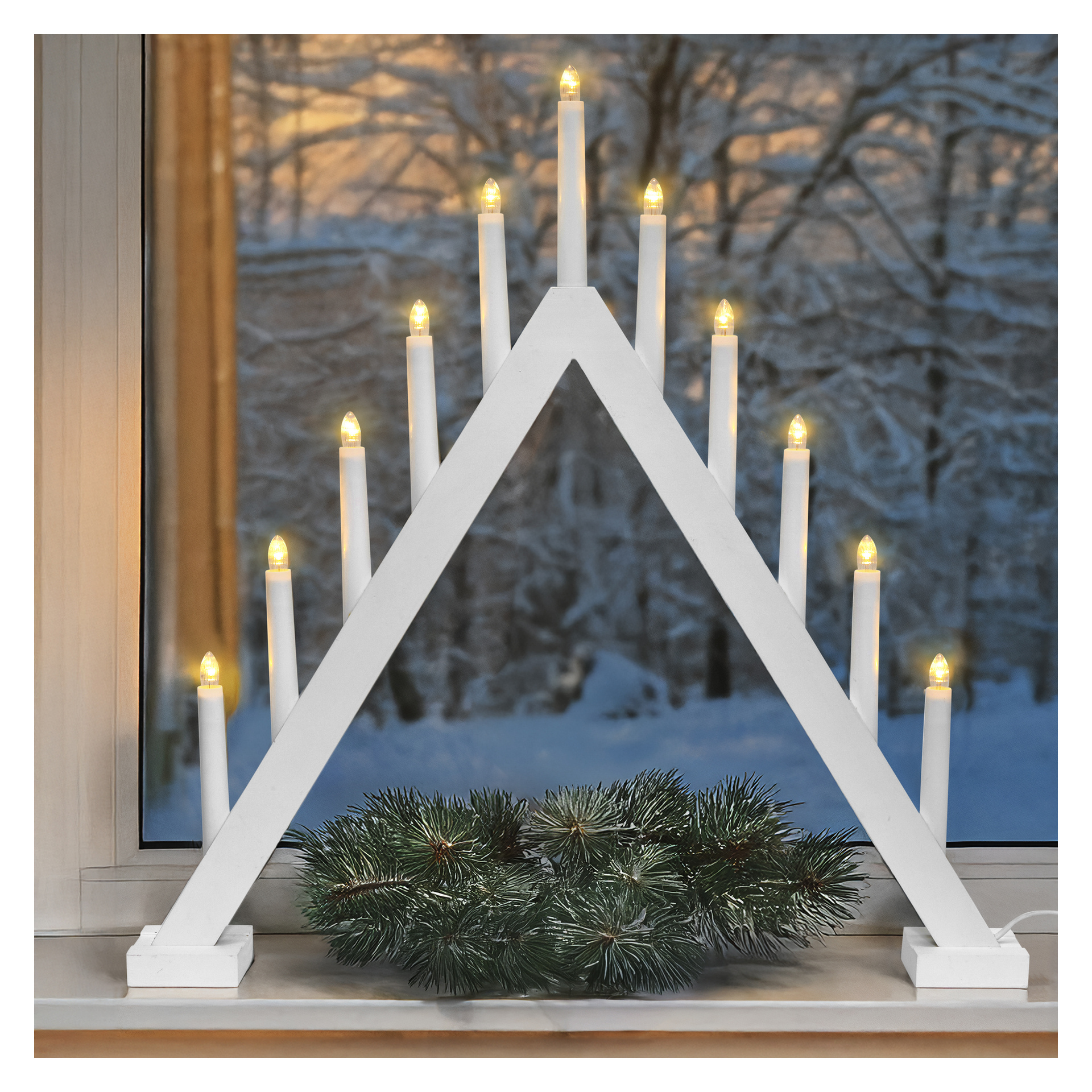 Der große LED-LED-Adventskerzenhalter EKVIP mit elf Lichtern in einem weißen Dreieck steht auf einer Fensterbank über Kiefernästen und verschneiten Bäumen.