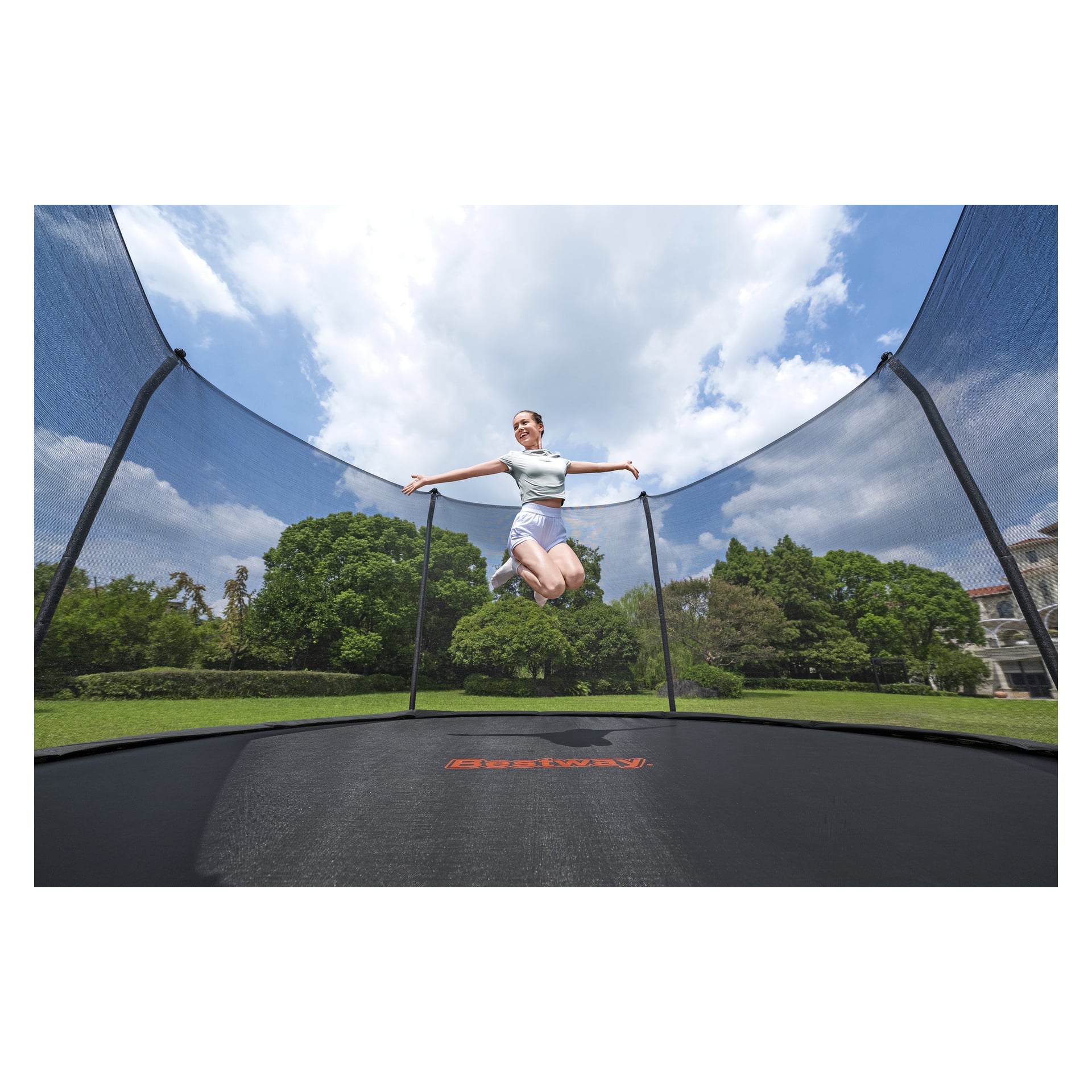 En person i sportsklær hopper på en Bestway Xtreme Air Trampoline 59120 med sikkerhetsnett, blant grønne trær og en bygning i sollys.
