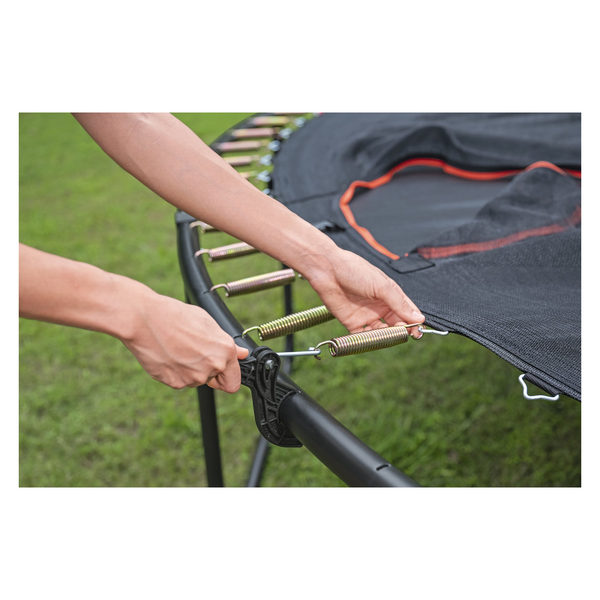 En person fester en metallfjær til rammen på en Bestway Xtreme Air 59120 trampoline, og fester den svarte matten over grønt gress.