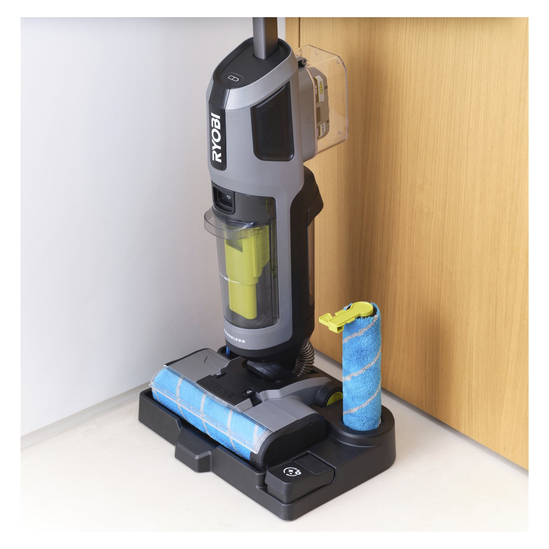 En Ryobi One+ 3-i-1 stående dammsugare med mopp, klar soptunna och blå rull står på sin laddare med träpanel; Extra rull i närheten.