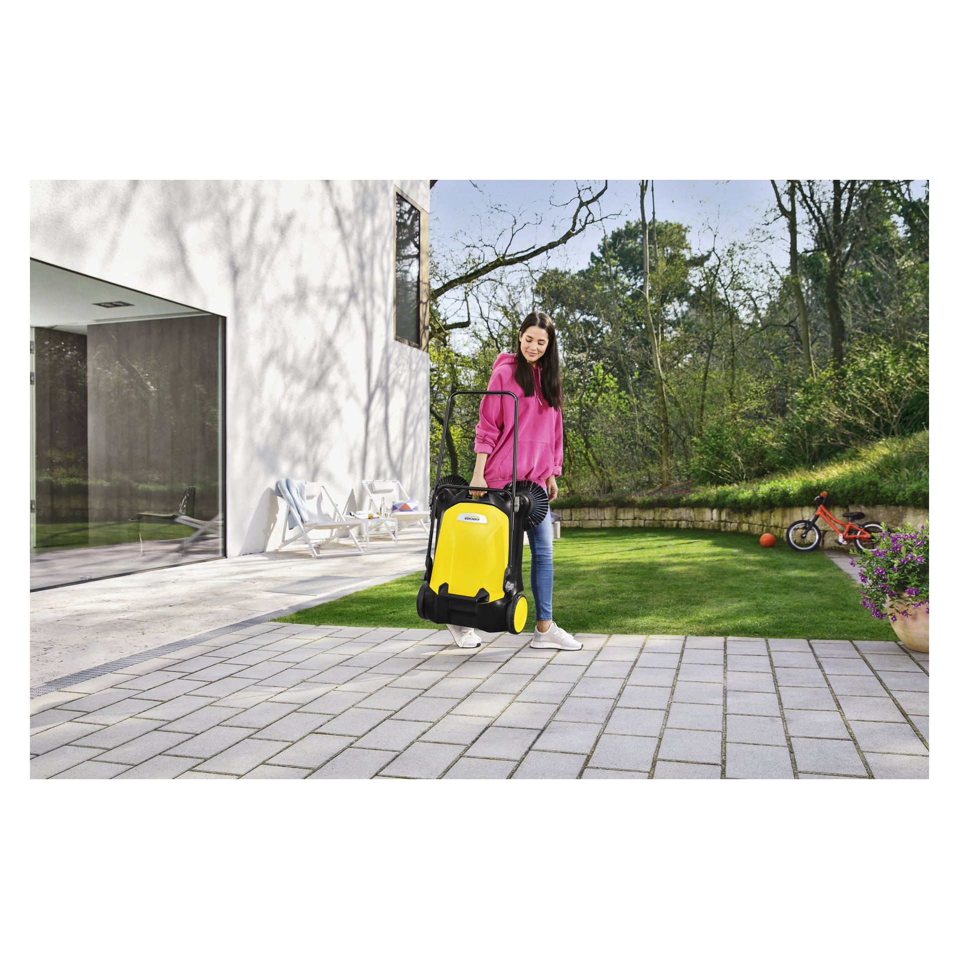 En kvinne i rosa hettegenser bruker en Kärcher S4 Twin sweeper på en terrasse ved et moderne hus, med gress, trær og en barnesykkel i nærheten.