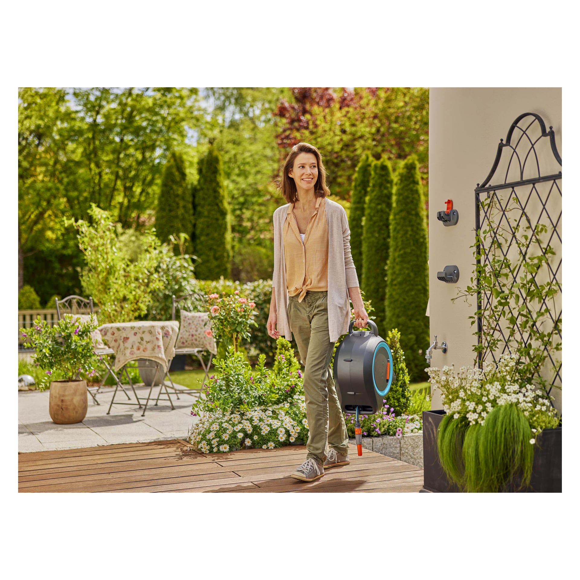 En kvinna på en träuteplats håller Gardena Hose Reel Dual M/L, med gröna växter, blommor och utemöbler i en solig trädgård.