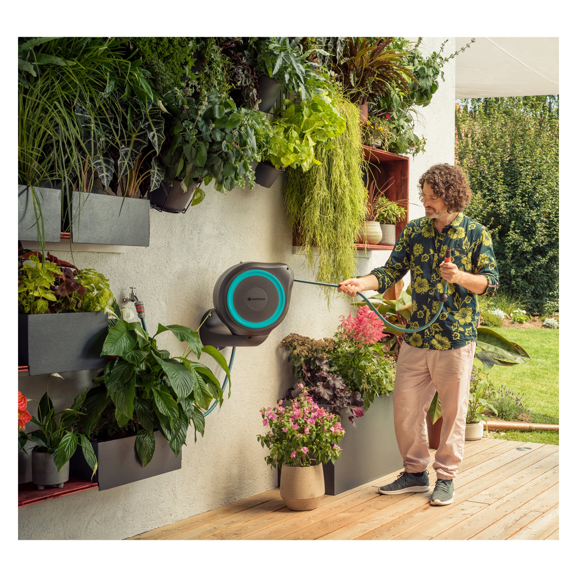 Eine Person zieht die Gardena Schlauchrolle RollUp M auf einer Holzterrasse, umgeben von verschiedenen Topf- und Wandbepflanzungen.