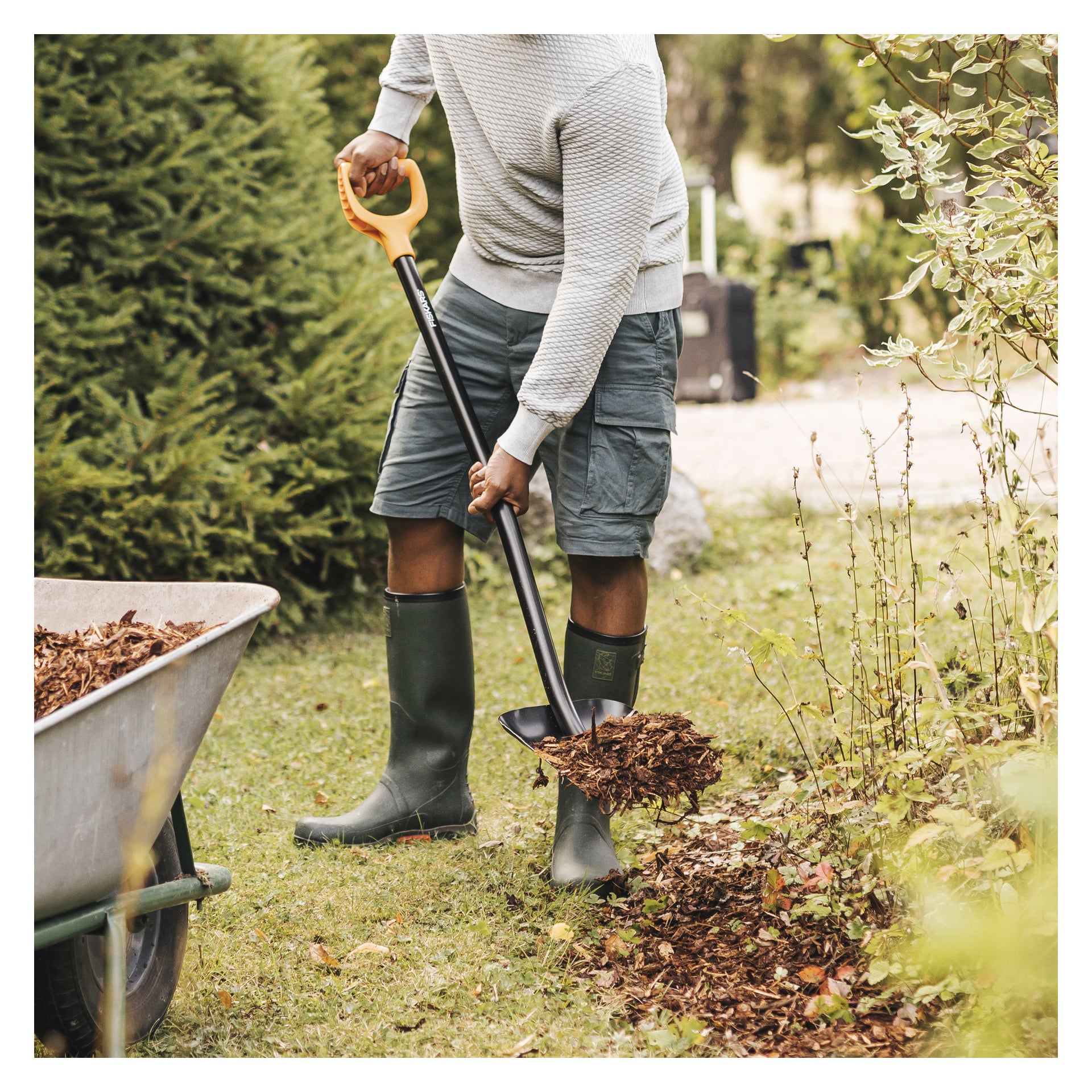 En person i en lett genser, shorts og gummistøvler smører bark fra en trillebår på en hagebed med en Fiskars Shovel Solid.