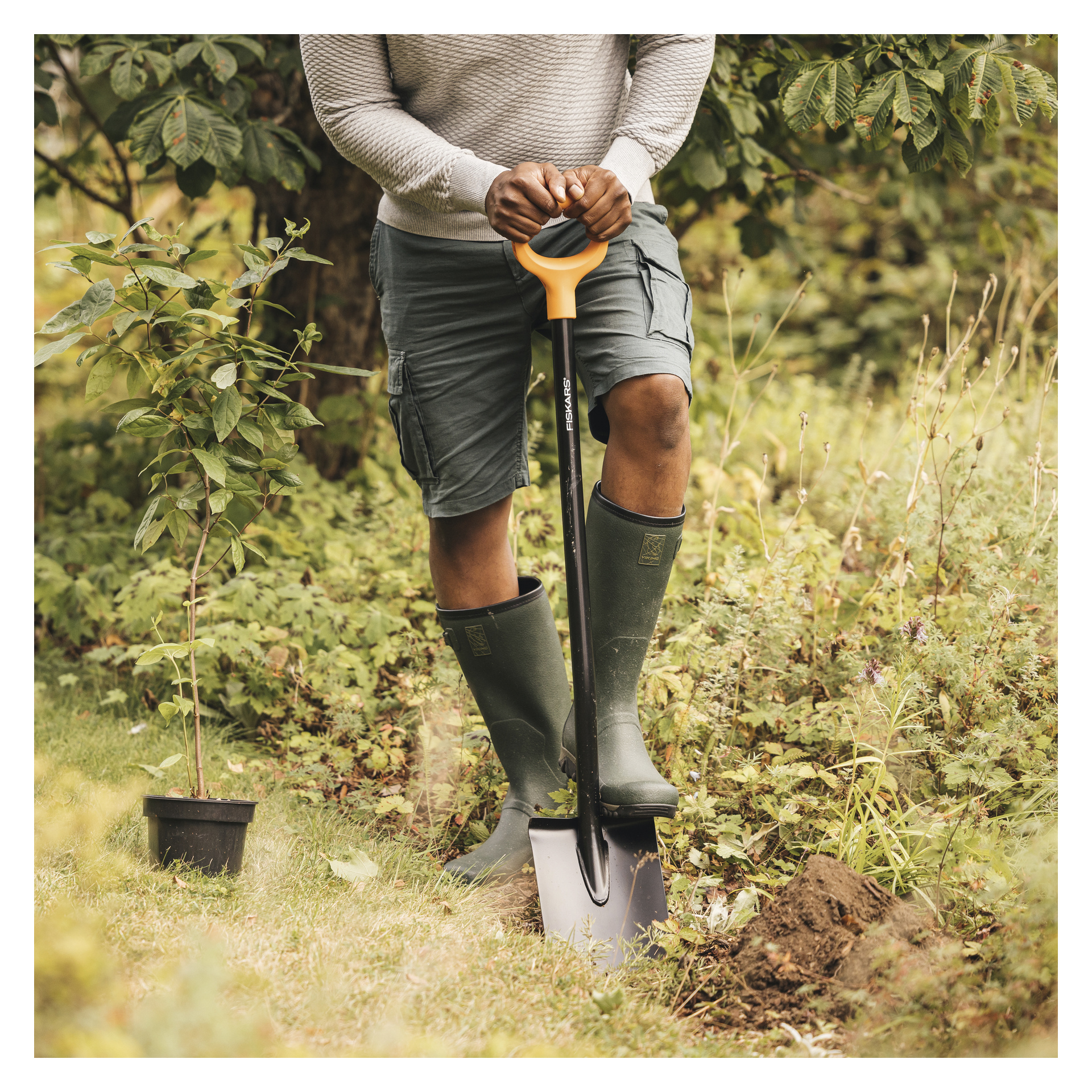 Iført en grå genser, grønne shorts og støvler, graver en person i en gresskledd hage med en Fiskars Pointed Spade Solid.