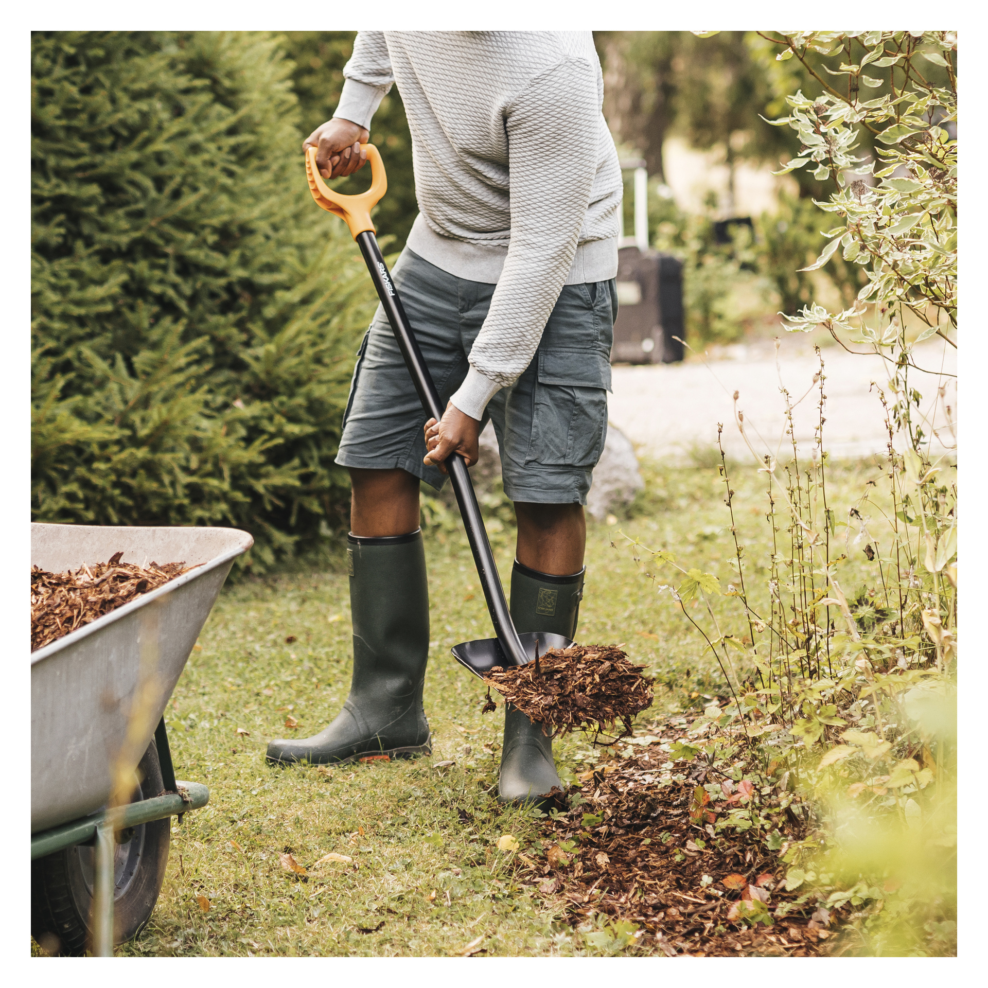 En person i genser, shorts og gummistøvler skyver mulch fra en trillebår ned på en hagebed med en Fiskars-spade.