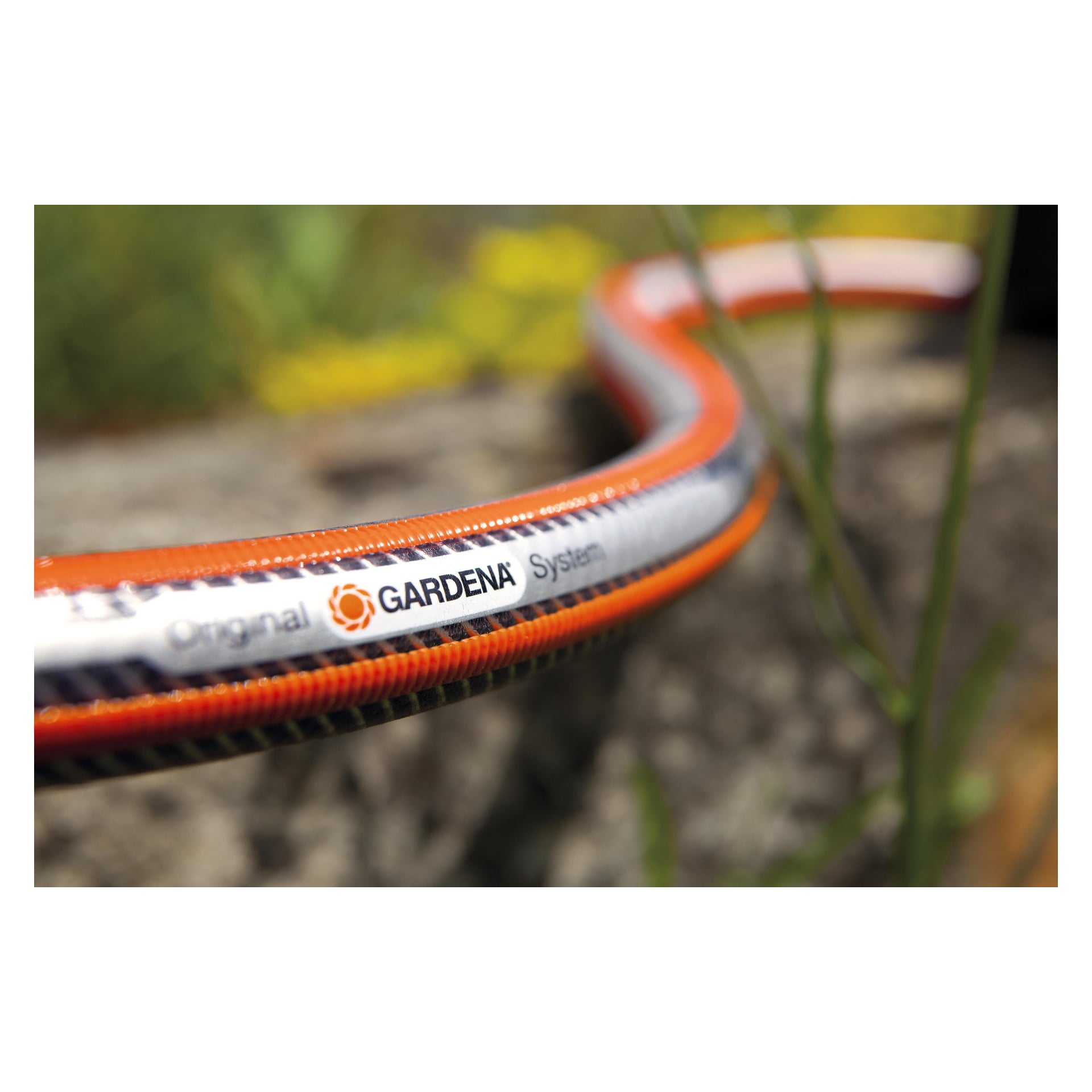 Närbild på Gardena Comfort HighFLEX-slangen med orange, vita och svarta ränder på sten bredvid gröna växter och gula blommor.