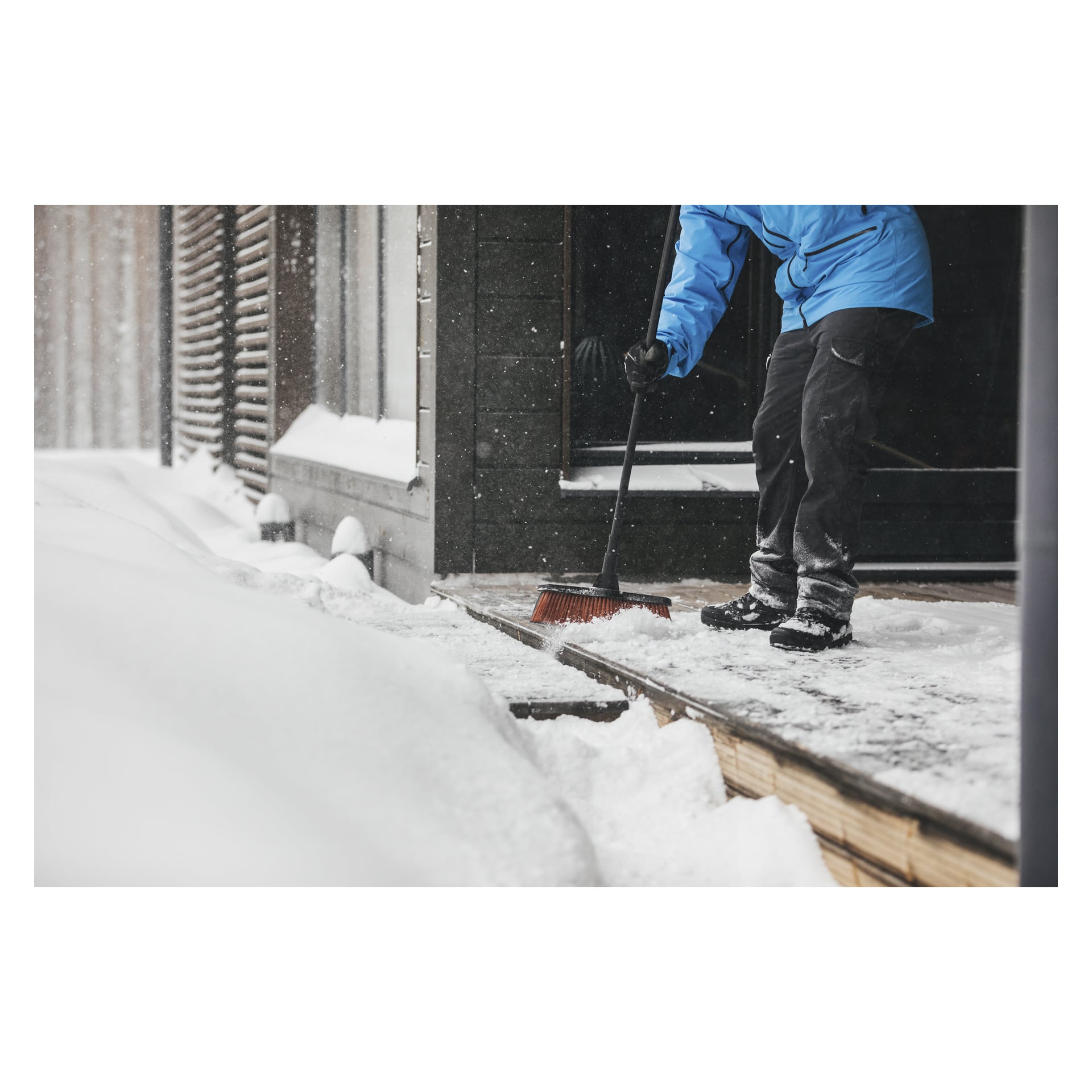 En person i blå jacka och svarta byxor sopar snö från en trägångväg med en Fiskars Broom Solid M medan snön faller runt.