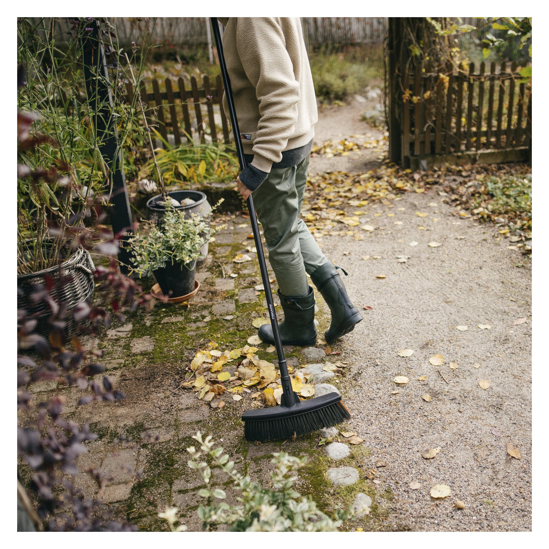 En person i stövlar, handskar och en beige tröja sopar löv längs en trädgårdsstig med en Fiskars Kvast Solid M nära krukväxter.