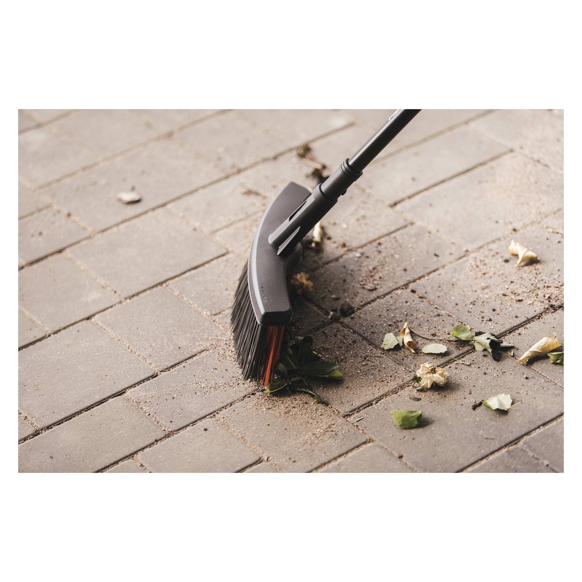 Närbild av en Fiskars Kvast Solid M som sveper damm, jord och löv från en belagd yta av rektangulära plattor.